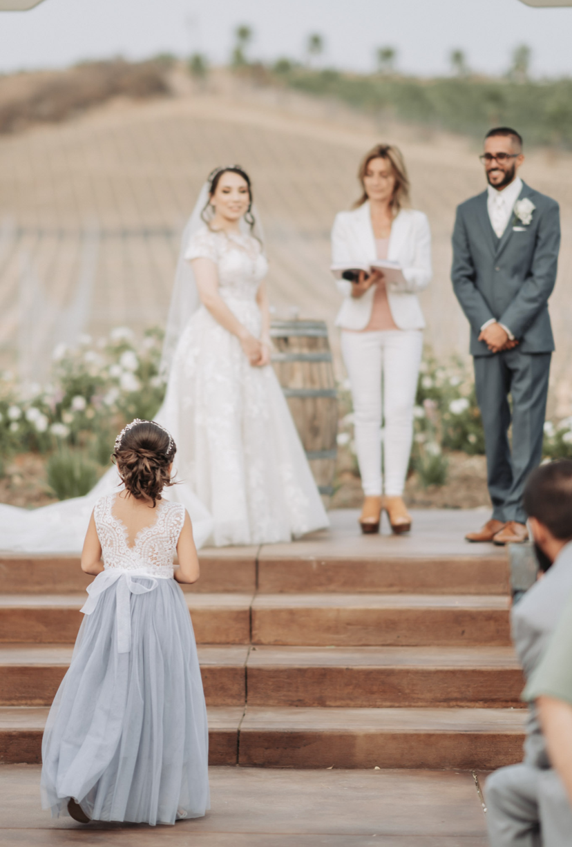 Two flower girls in pink dresses walk down an aisle at an outdoor wedding.