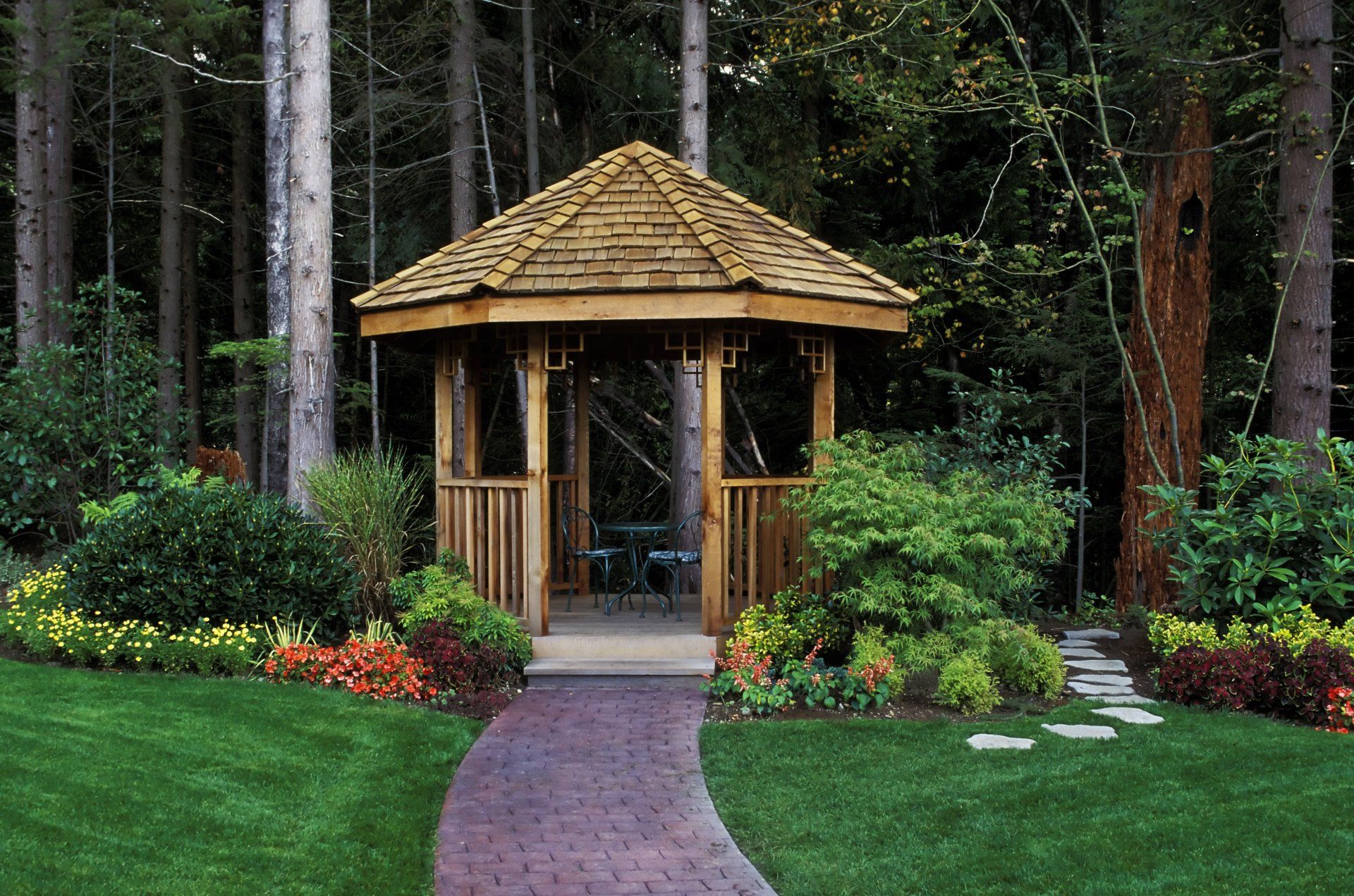 Gazebo da giardino con tetto di paglia, circondato da fiori e alberi, con un sentiero in pietra che conduce all'ingresso.