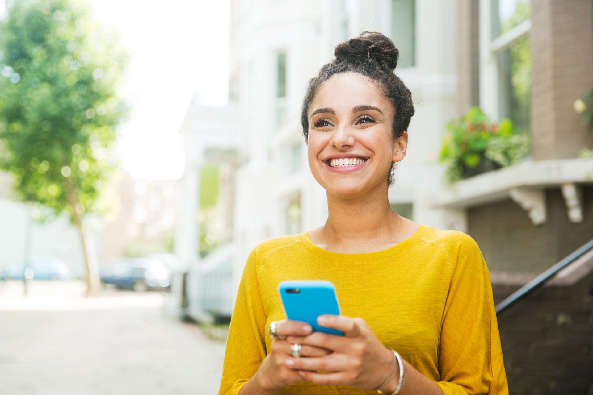 Persona sorridente con un maglione giallo che tiene in mano uno smartphone blu all'aperto in una strada soleggiata