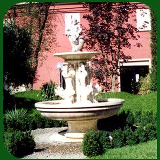 Fontana in pietra in un cortile con giardino antistante un edificio rosa, circondata dal verde.