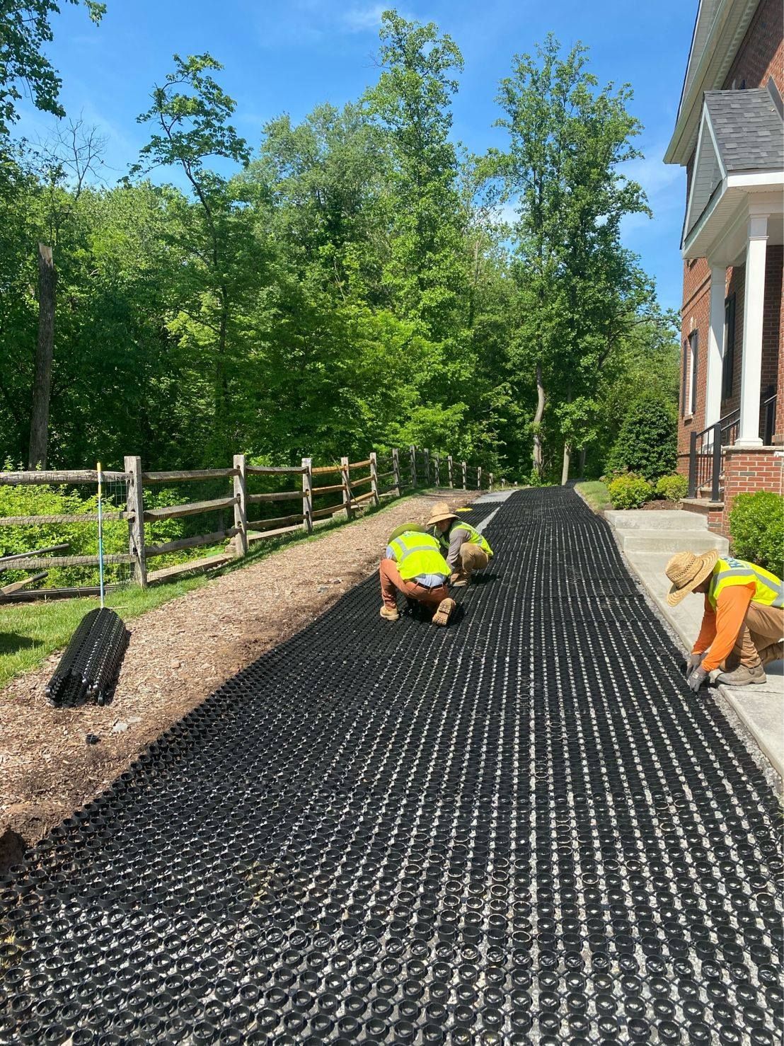 Workers installing black grid over a driveway, with gravel and a fence on the side and a house to the right.