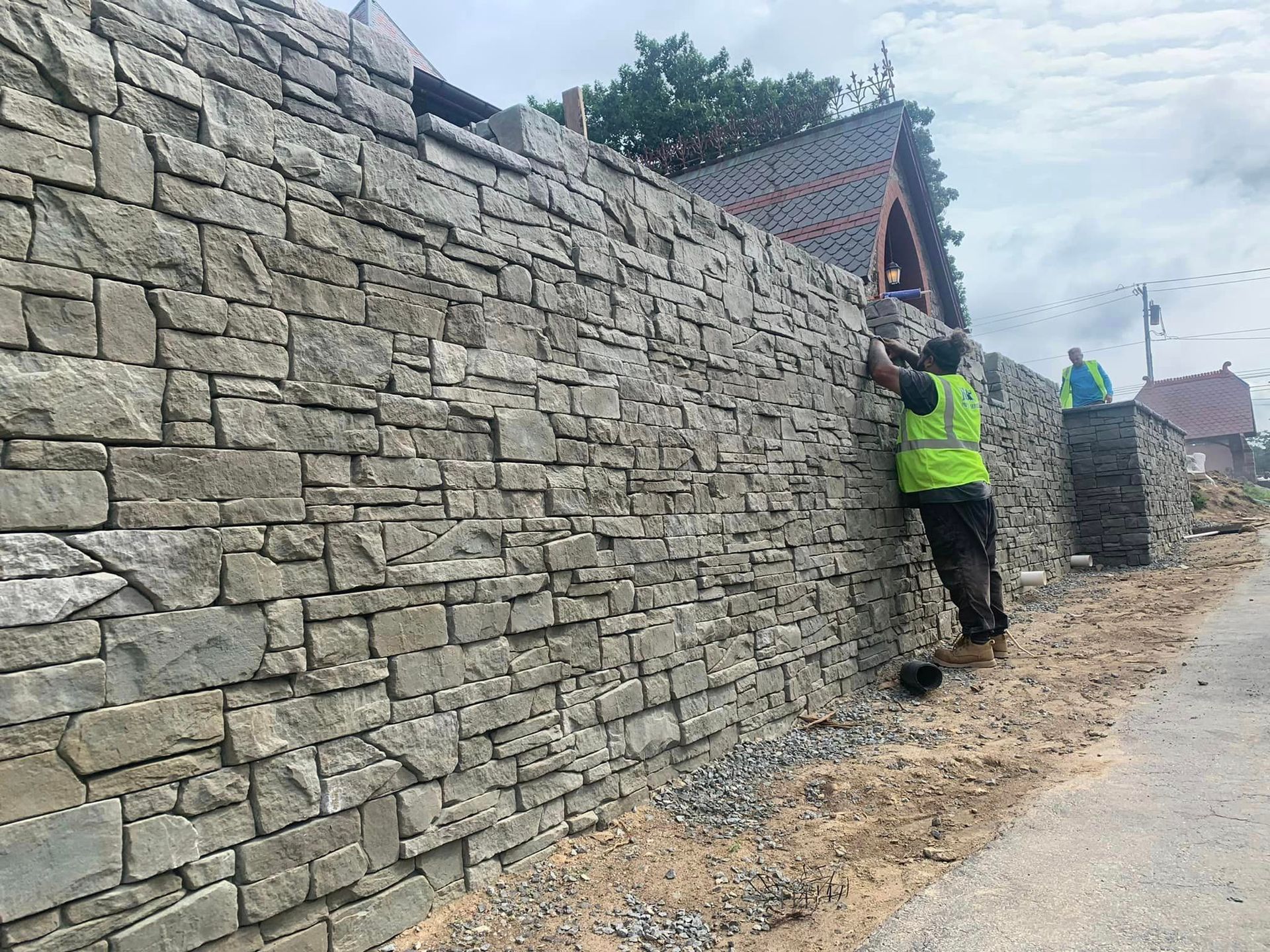Construction workers installing stone facing on a wall outdoors. One worker wears a safety vest.