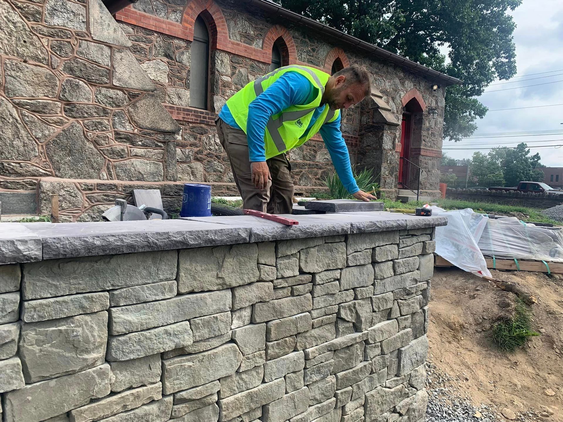 Man in safety vest works on stone wall, next to building with arched windows, outdoors.
