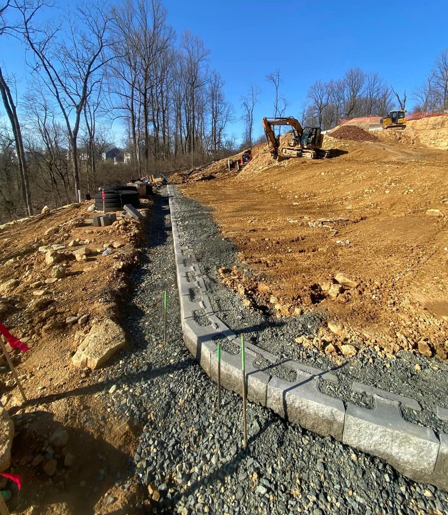 A retaining wall of concrete blocks under construction on a hillside with an excavator.
