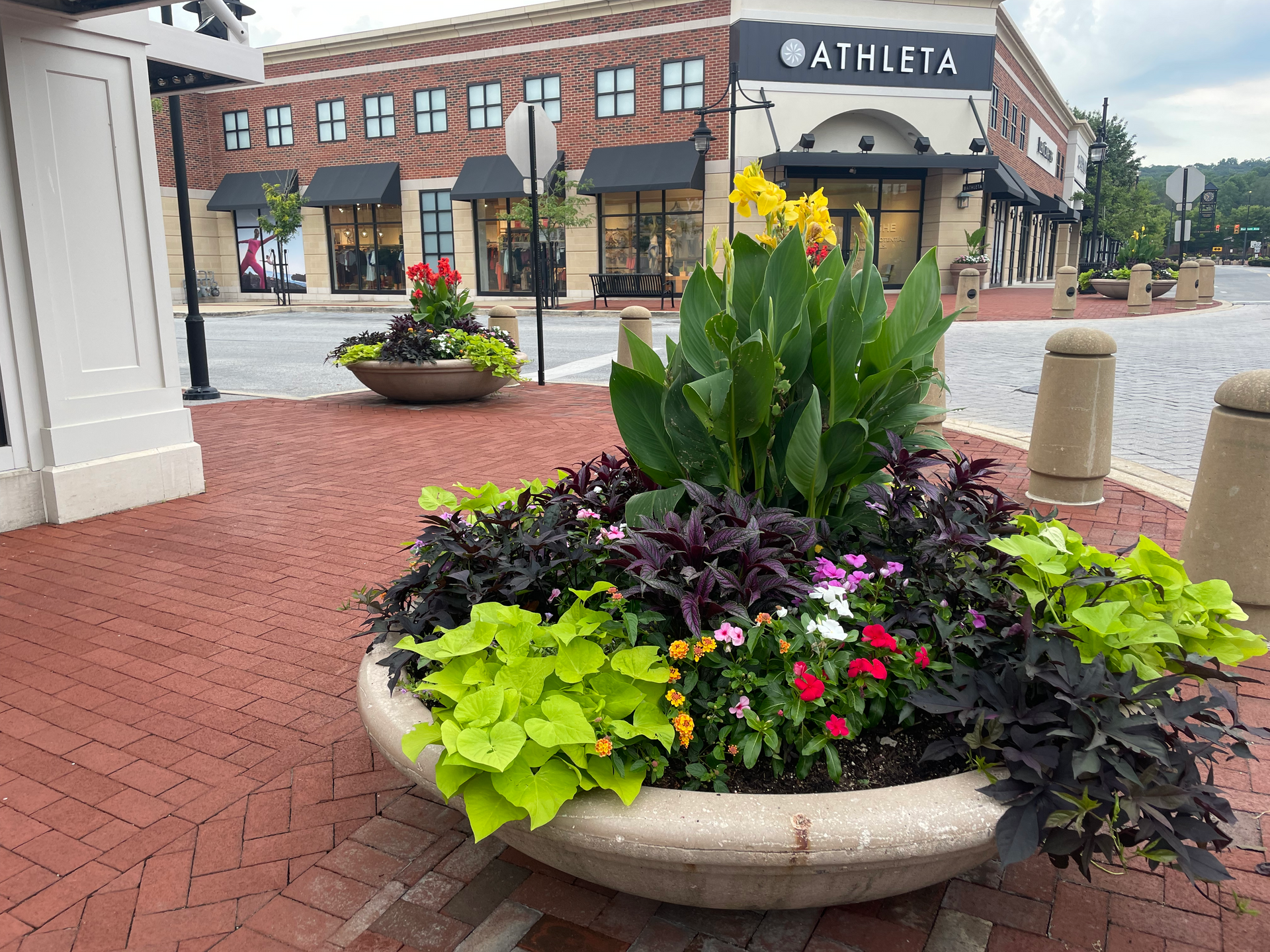 Flower planters on a brick walkway in front of Athleta store, with other stores and buildings in the background.