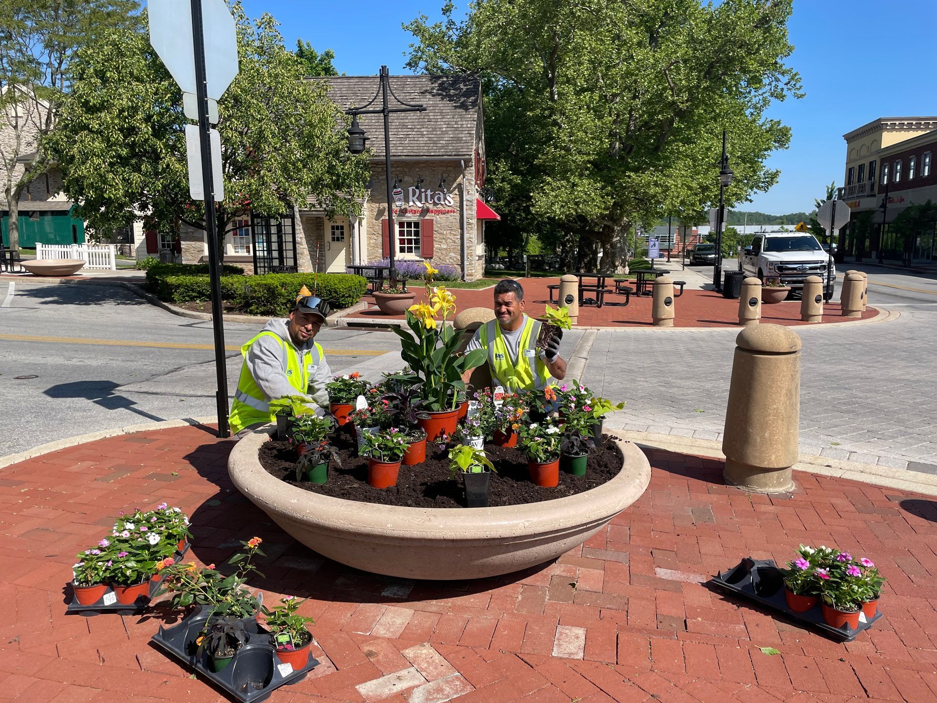 Two people in vests planting flowers in a large, round planter in a brick-paved town square.