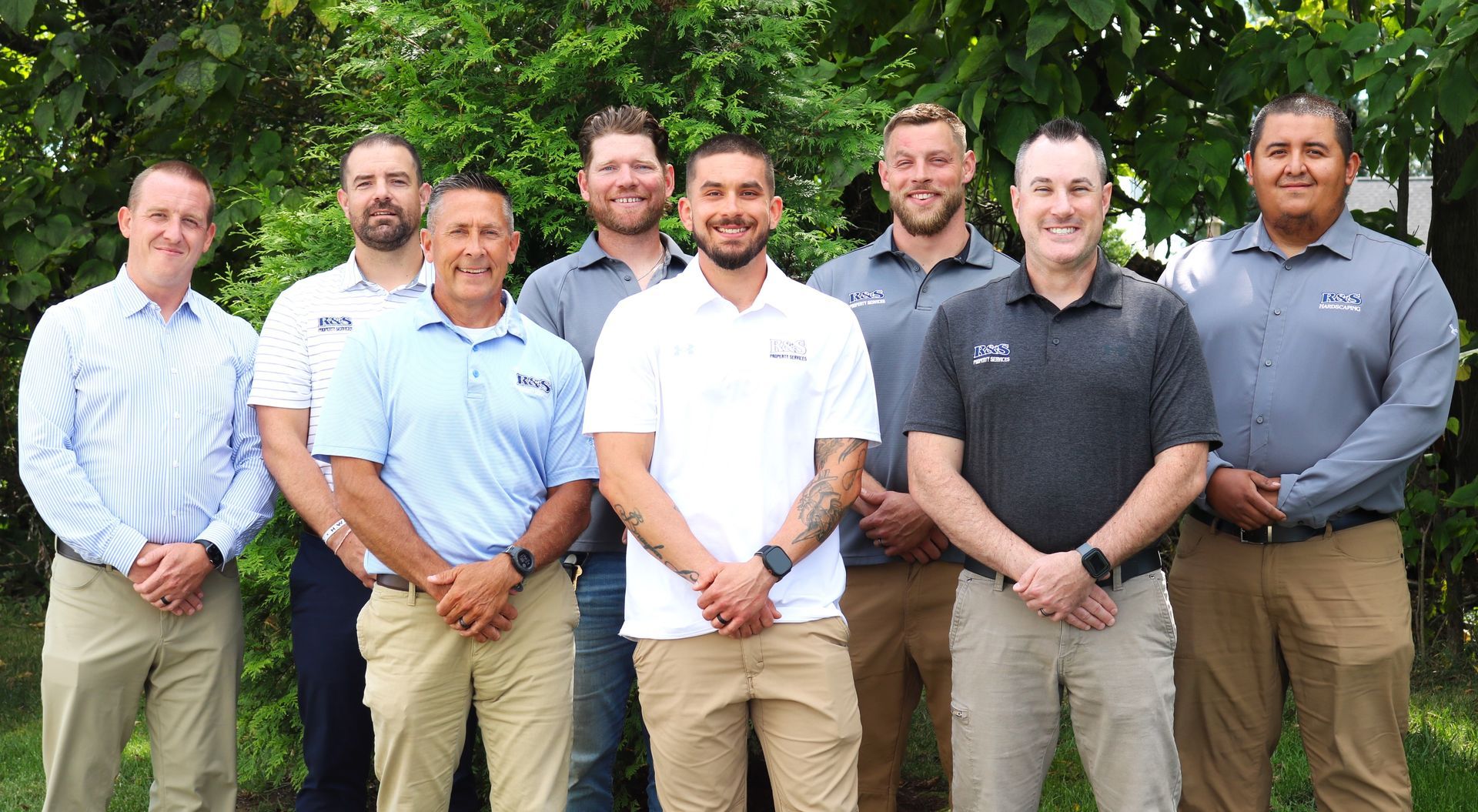 Group of nine men standing outdoors, smiling. They wear business casual attire, some with logos.