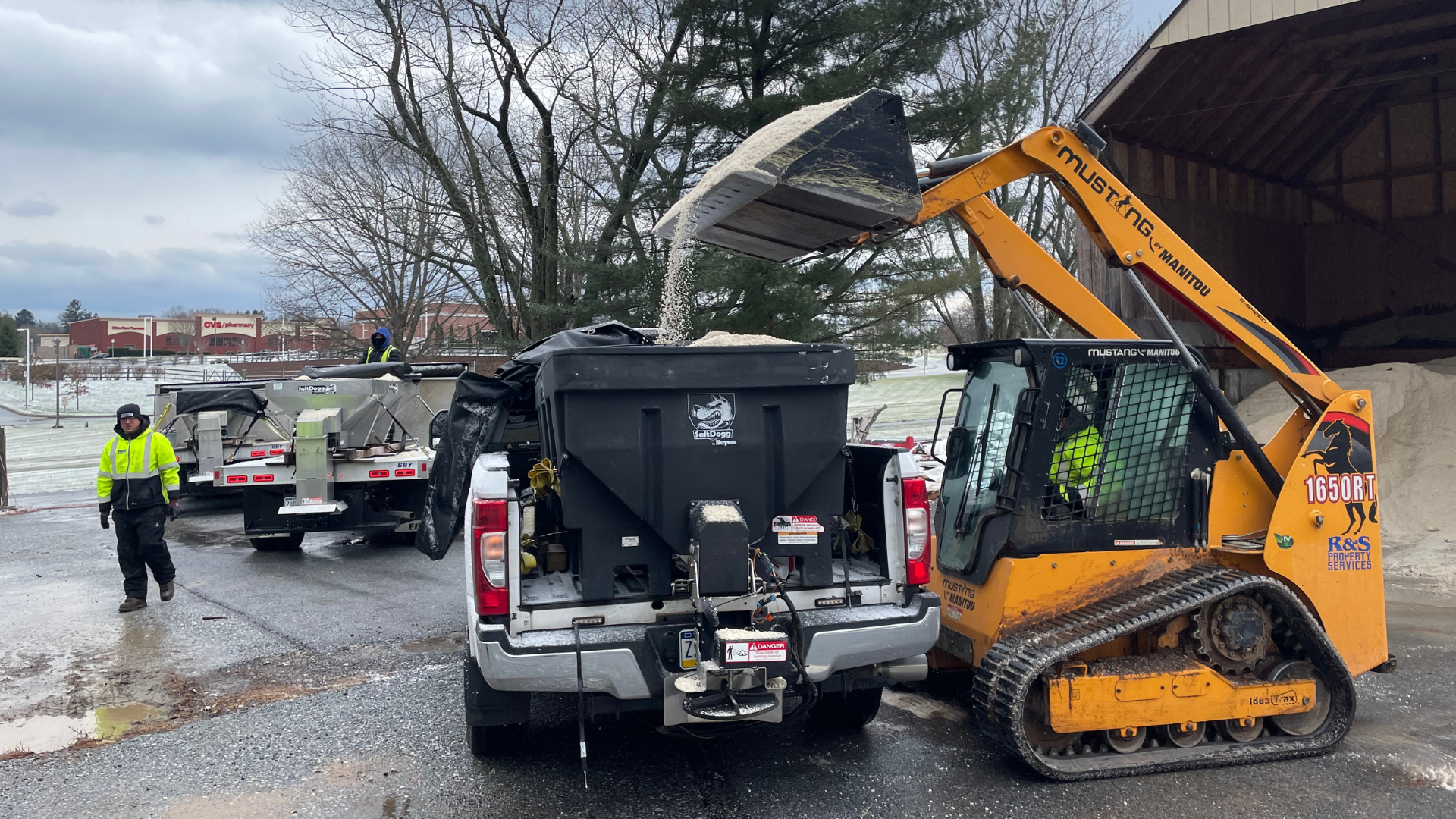 A skid steer loads salt into a pickup truck salt spreader. A worker stands nearby in a winter coat.