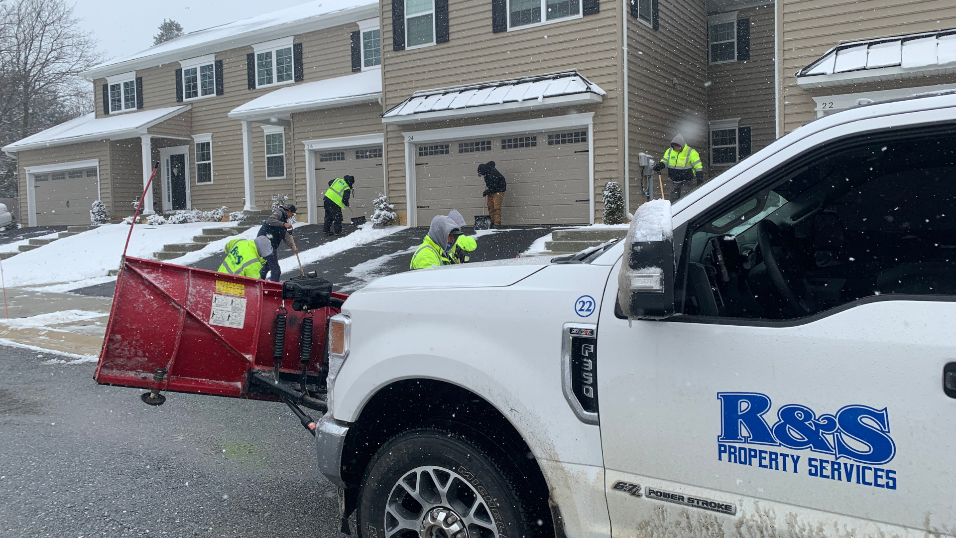 White truck with a plow clearing snow from a driveway, people shoveling snow near houses.