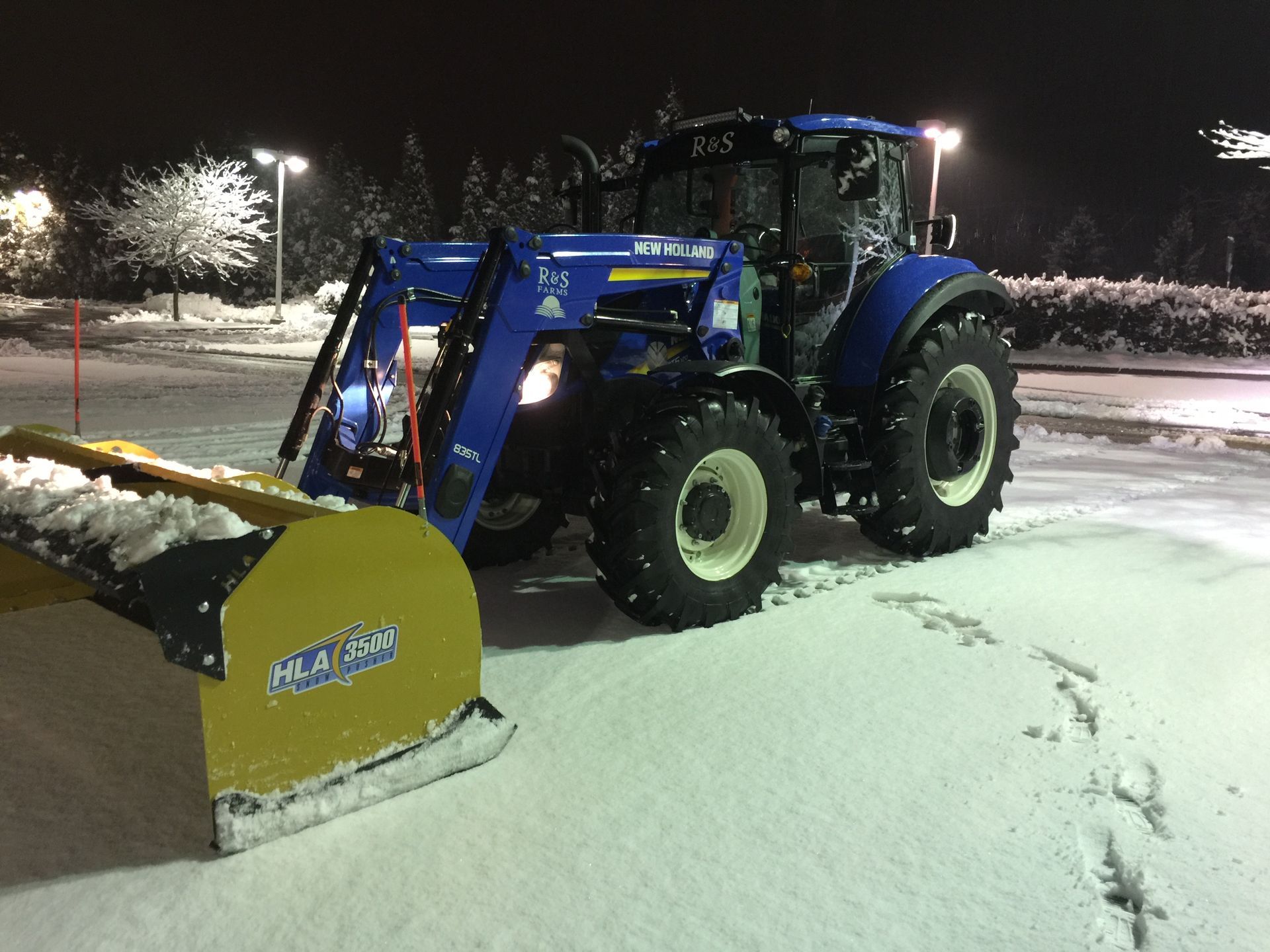 Blue tractor plowing snow in a parking lot at night, with a yellow snowplow blade.