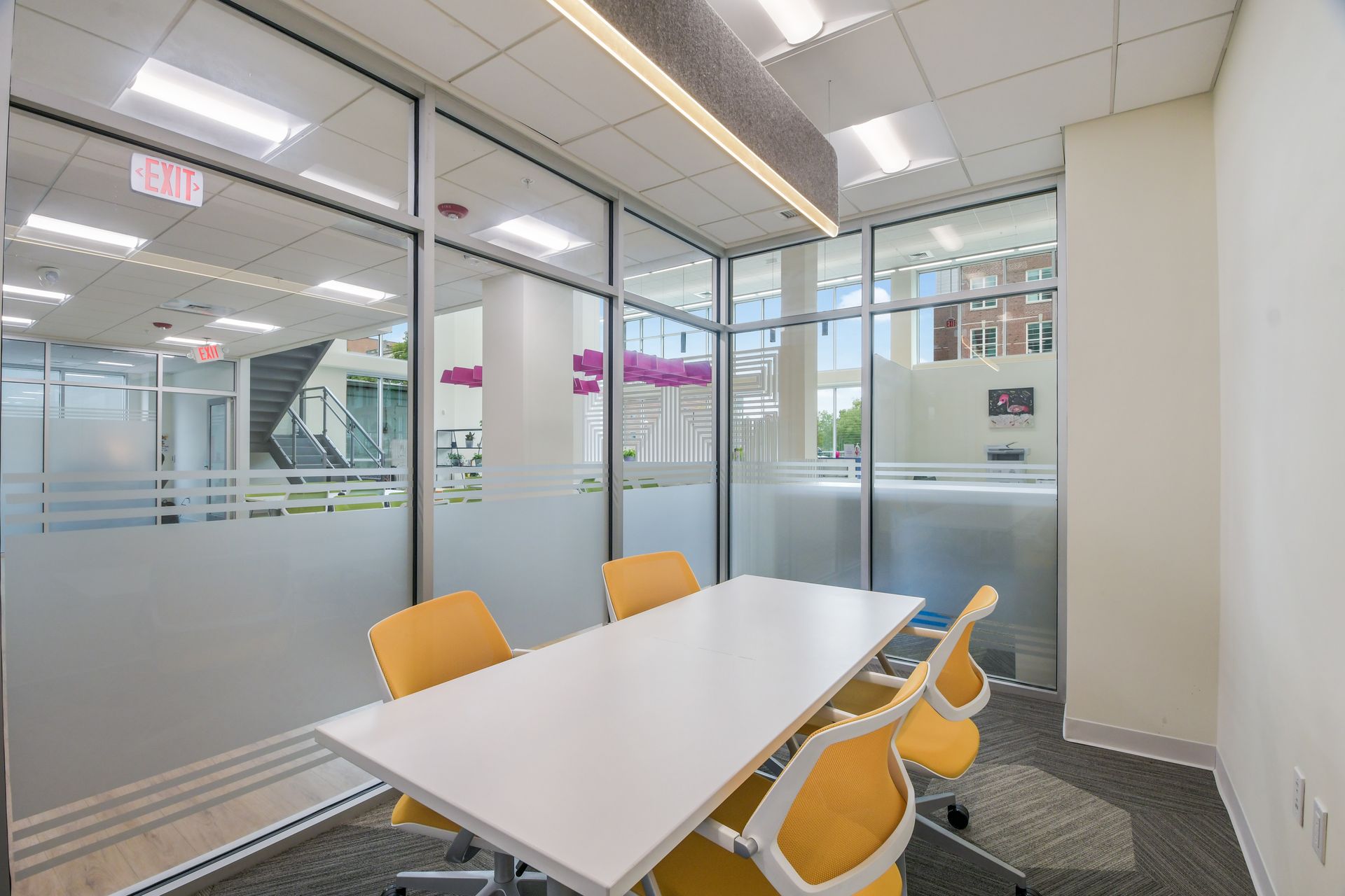 A small, white-tabled meeting room with yellow chairs, glass walls, and a view of a larger office space.