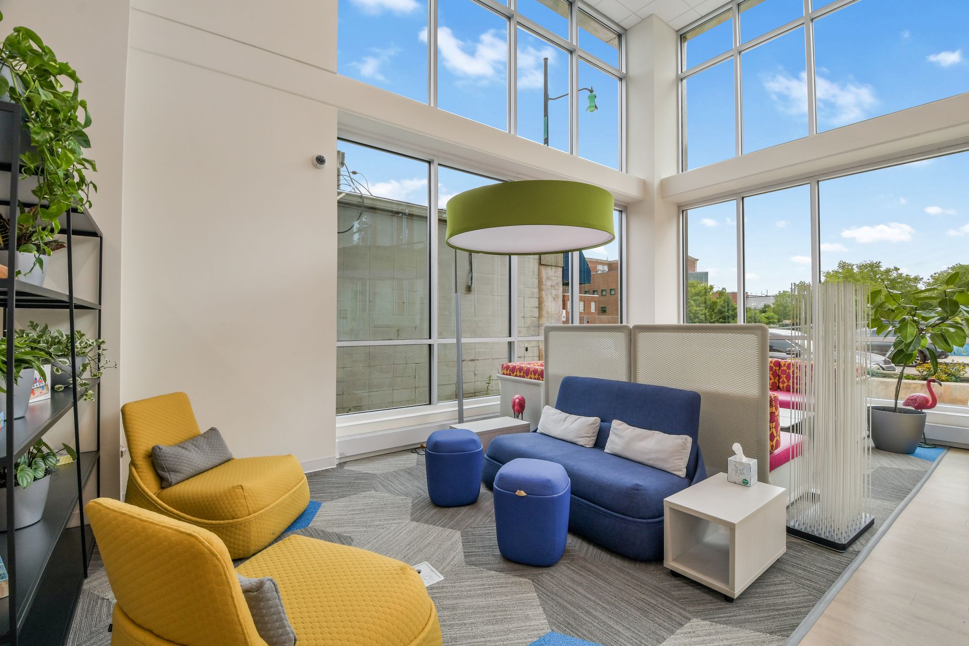 Lounge area with blue sofa, yellow chairs, and large windows. Includes plants and modern decor.