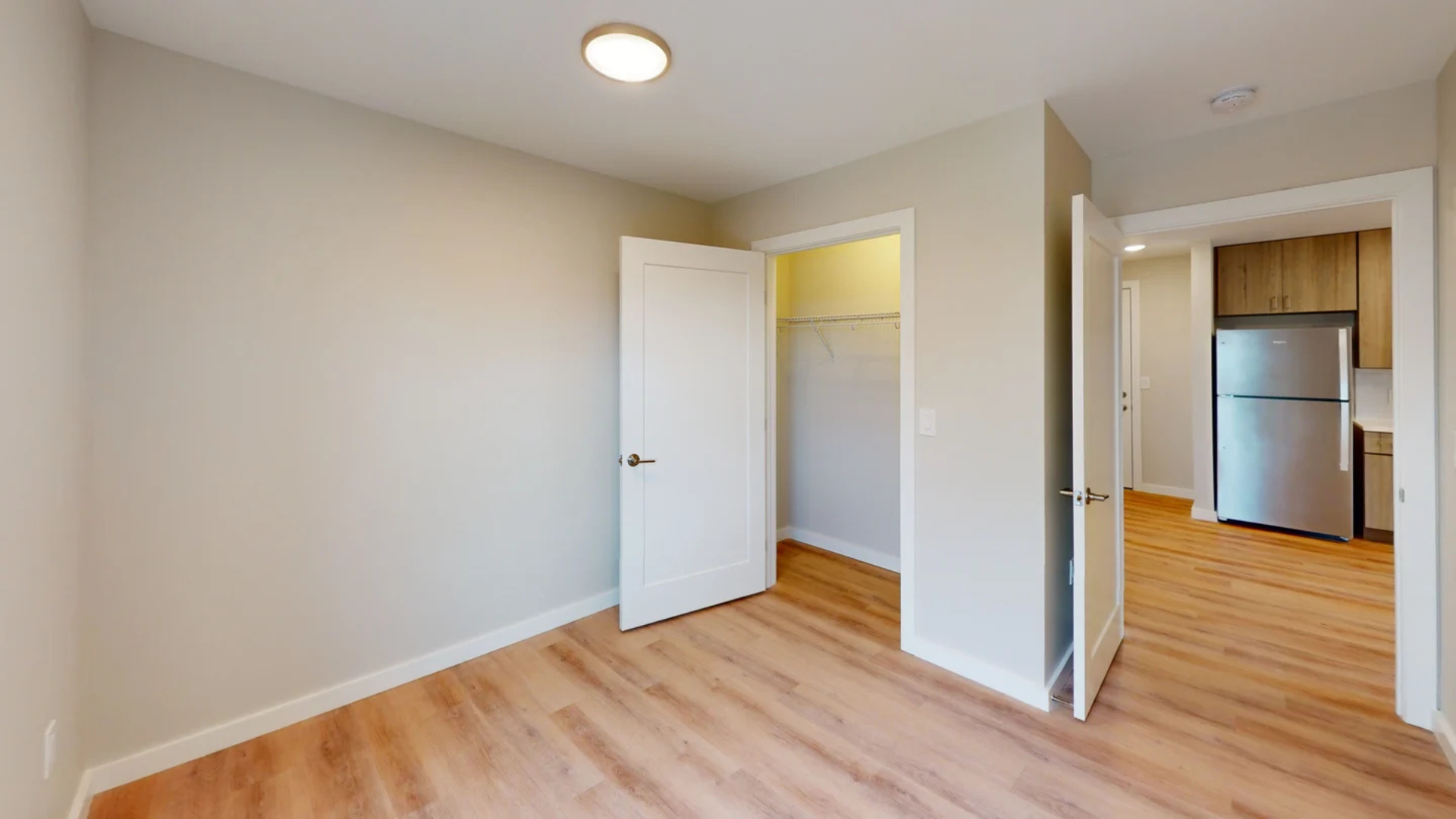 Bedroom interior with open doors to a closet and kitchen, light wood flooring, and beige walls.