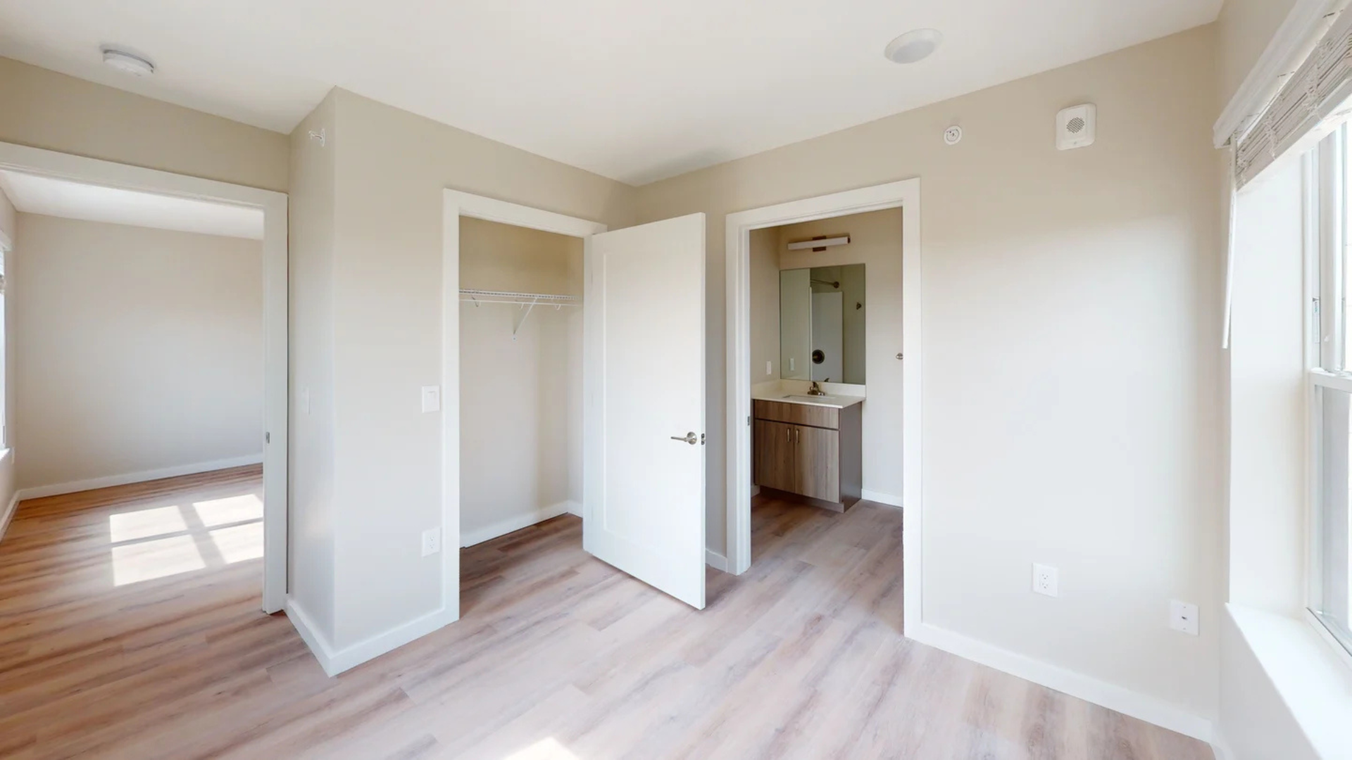 Empty bedroom with closet and doorway to bathroom, light wood floors, neutral walls, and a window.