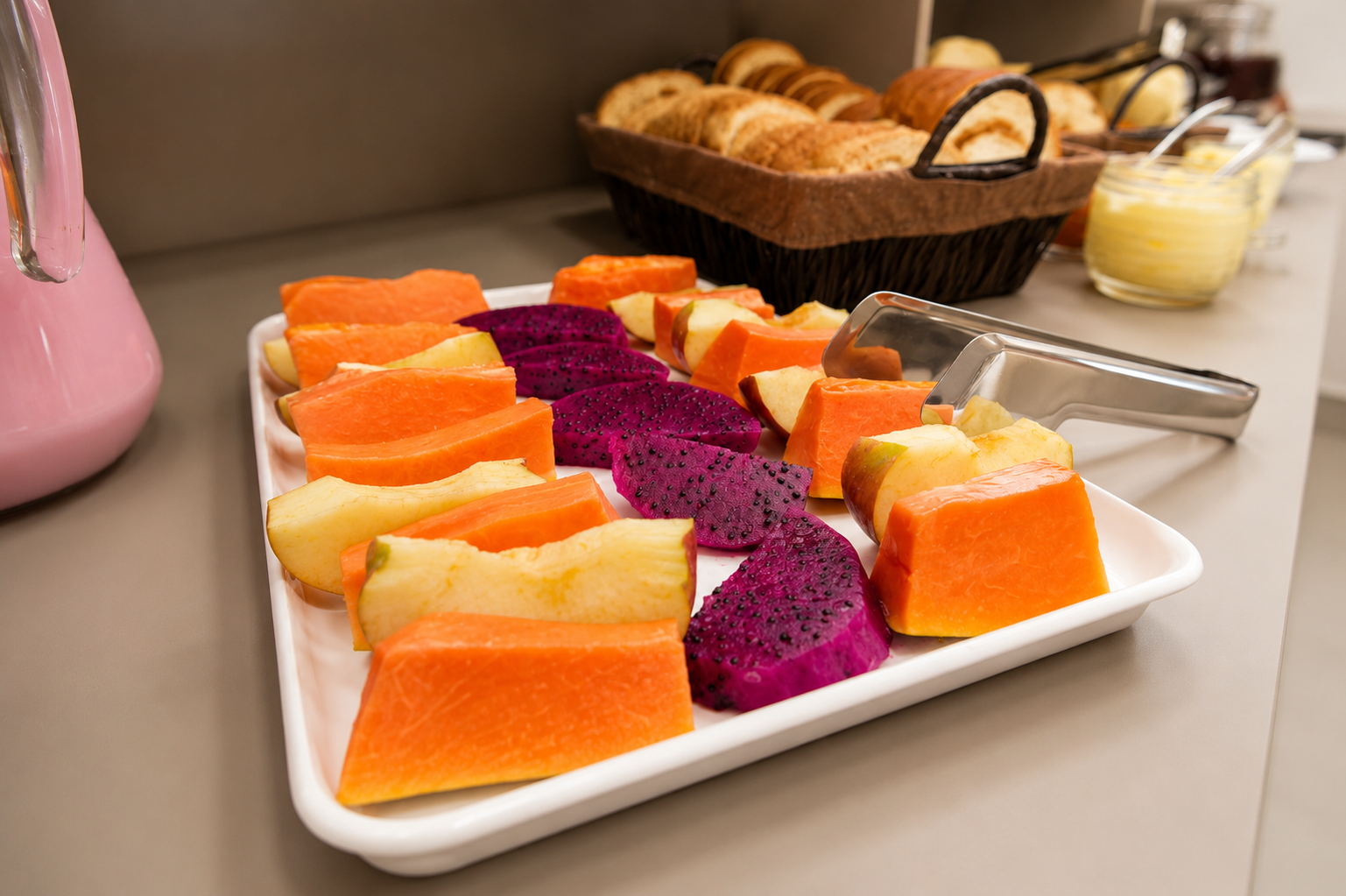 Fatias de frutas coloridas variadas dispostas em um prato branco com pão e manteiga ao fundo.