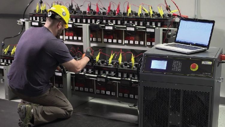 Técnico con casco amarillo trabajando en una fila de bastidores de baterías junto a un ordenador portátil y una unidad de control.