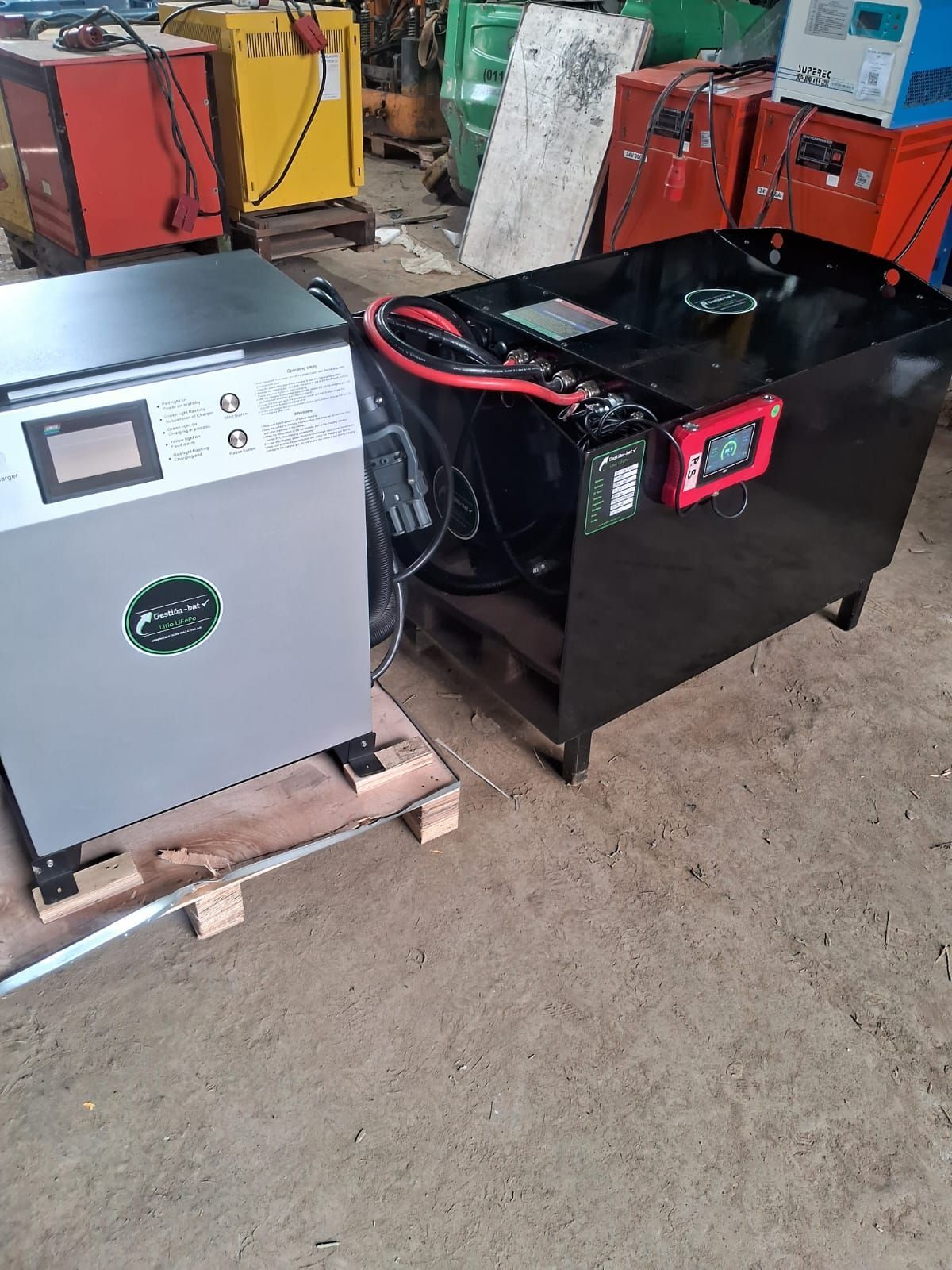 Dos unidades de baterías industriales sobre palés, una gris y otra negra con cables rojos, en un almacén.