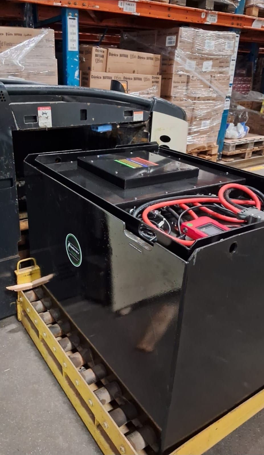Unidad de batería industrial negra sobre un palé de almacén con cables rojos y cajas de cartón al fondo.