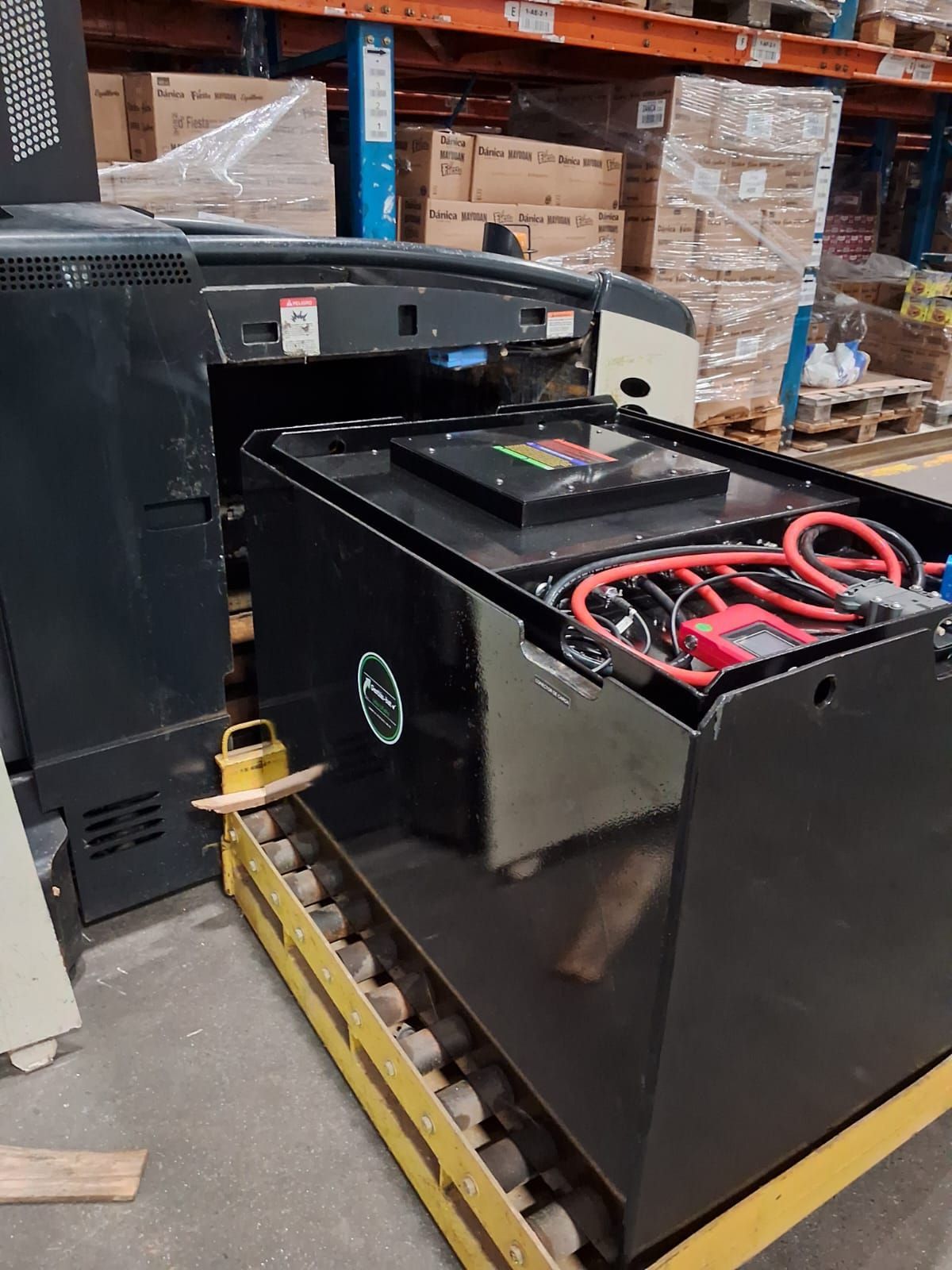 Batería negra para carretilla elevadora sobre un palé en un almacén, con cables rojos y cajas apiladas cerca.