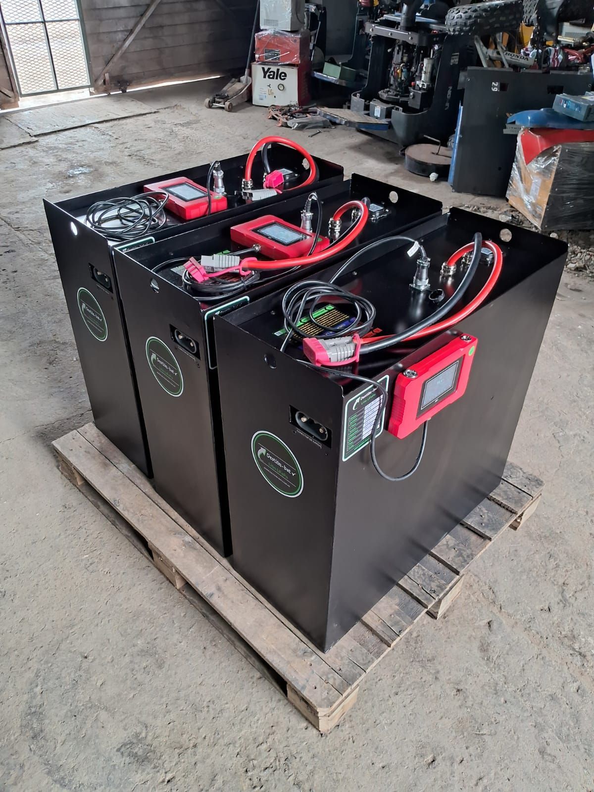 Tres paquetes de baterías negras con cables rojos sobre un palé de madera en un taller industrial.