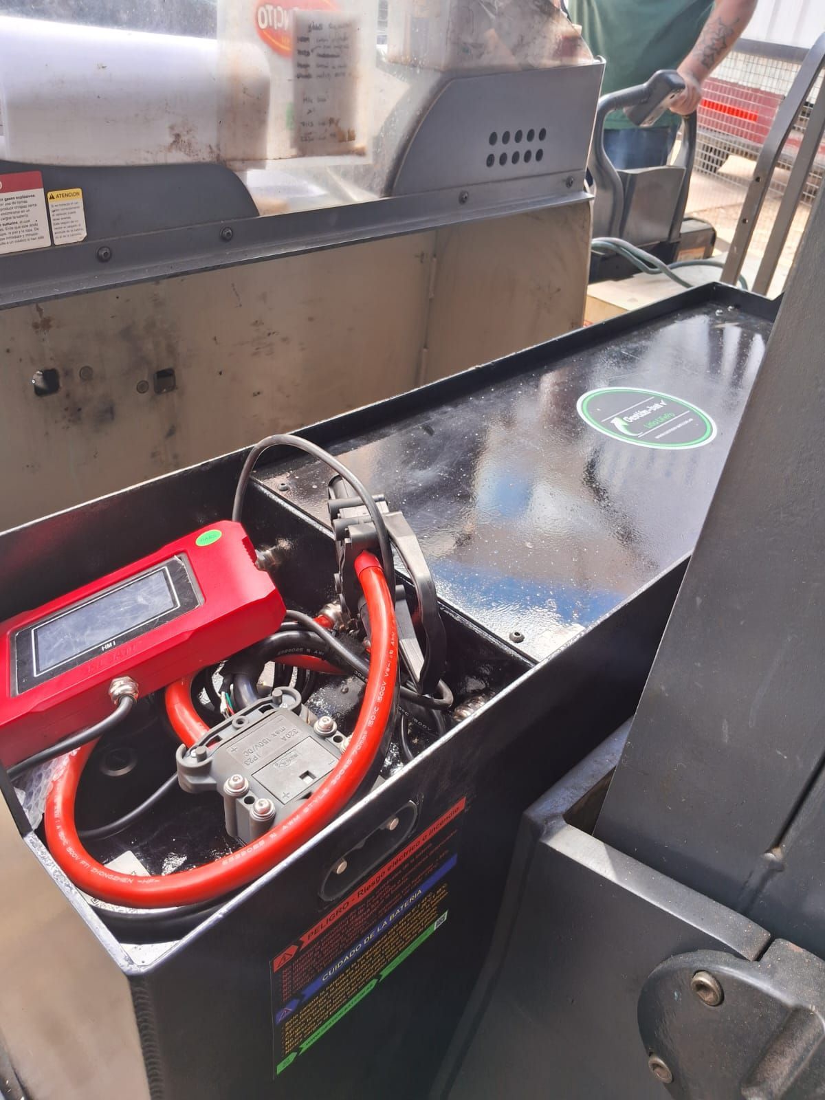 Batería portátil roja con cables en una caja de herramientas negra junto a una parrilla o ahumador al aire libre.