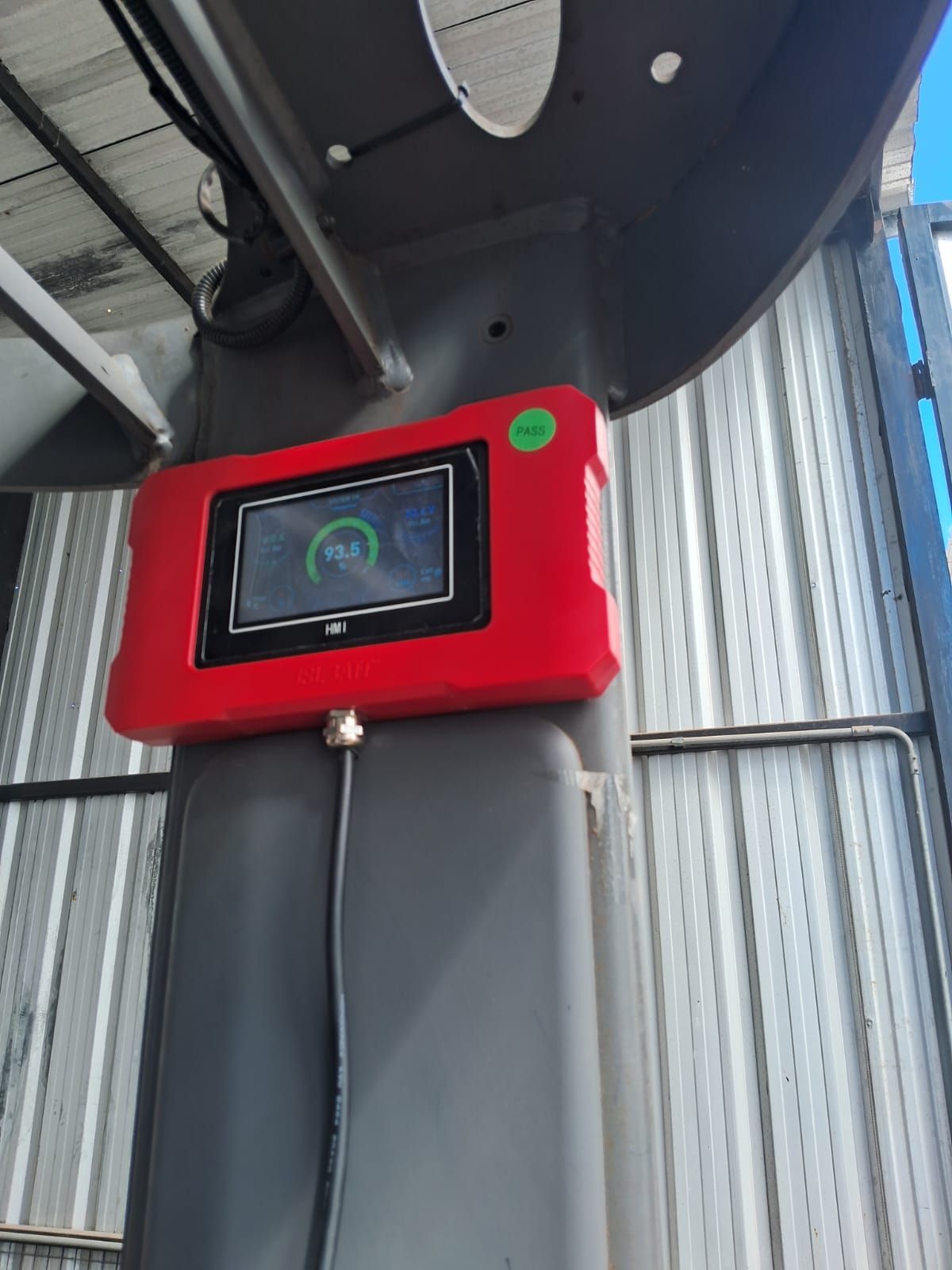 Caja de control roja montada en la pared con una pequeña pantalla bajo un techo metálico en un entorno industrial.