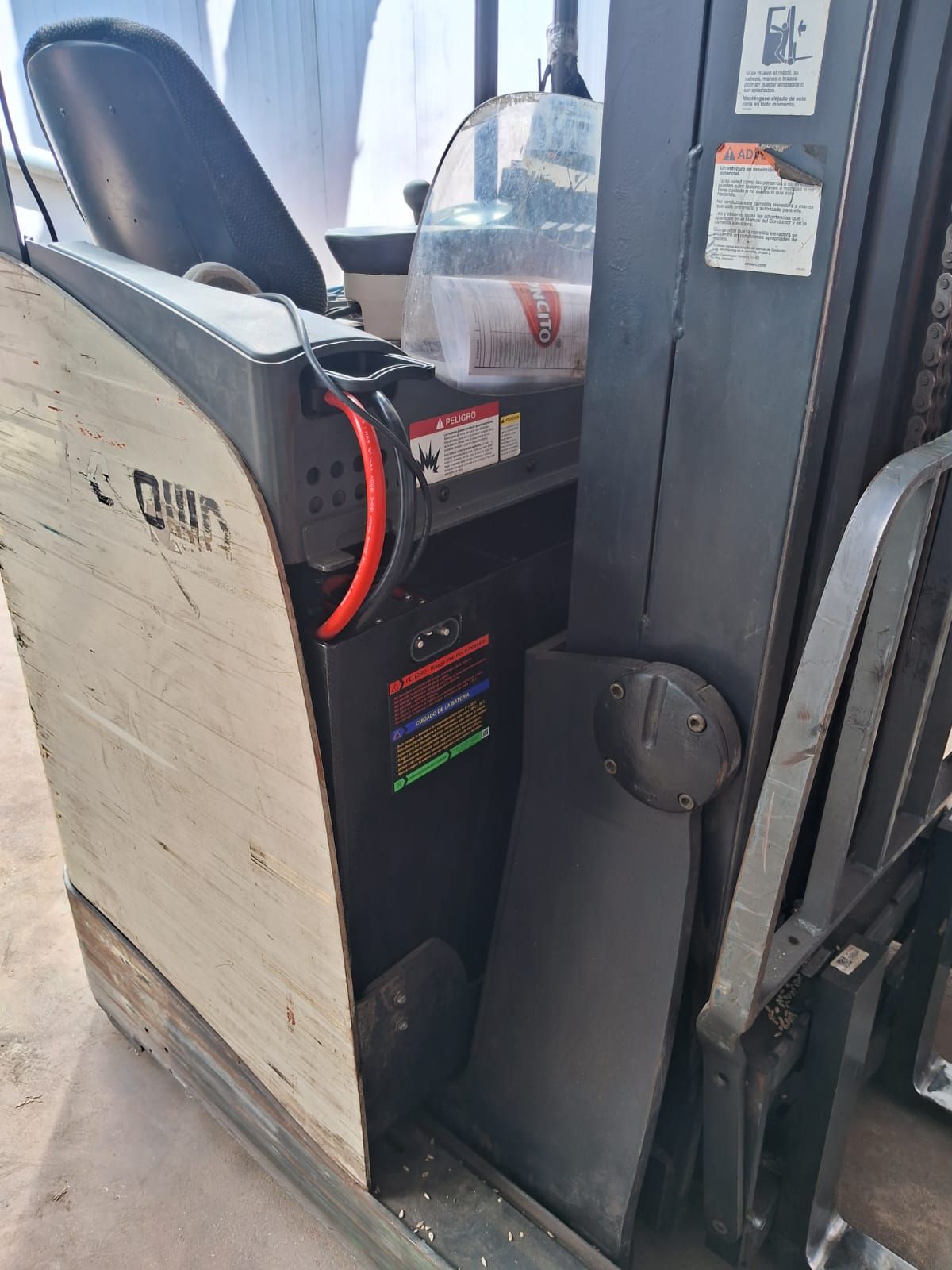 Asiento del operador de la carretilla elevadora junto al mástil de una transpaleta, con una mampara de seguridad de plástico transparente y etiquetas de advertencia.