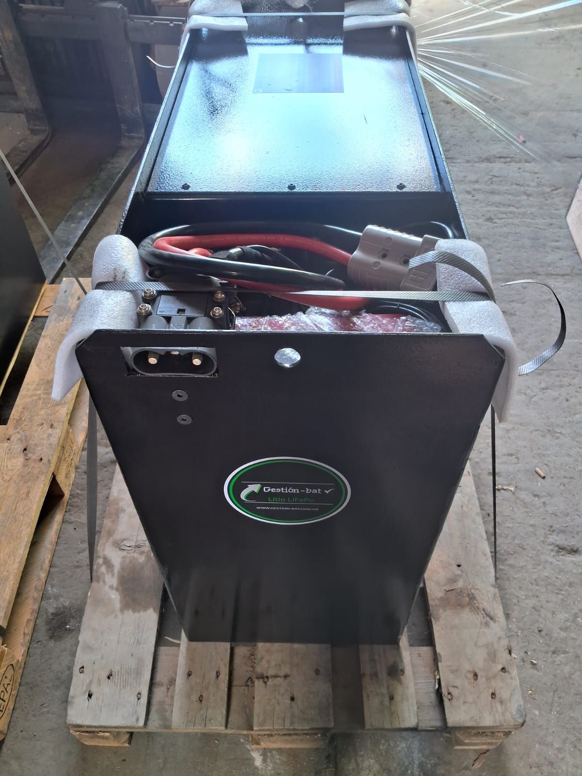 Cargador de baterías portátil negro sobre un palé de madera con cables rojos y negros conectados.