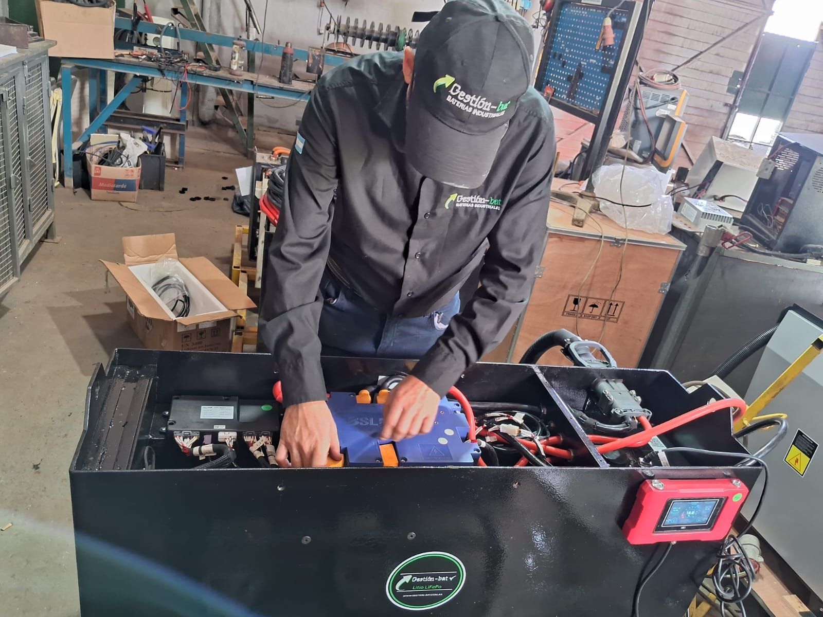 Un trabajador con gorro y chaqueta negros está cableando un dispositivo azul dentro de una máquina negra en un taller.