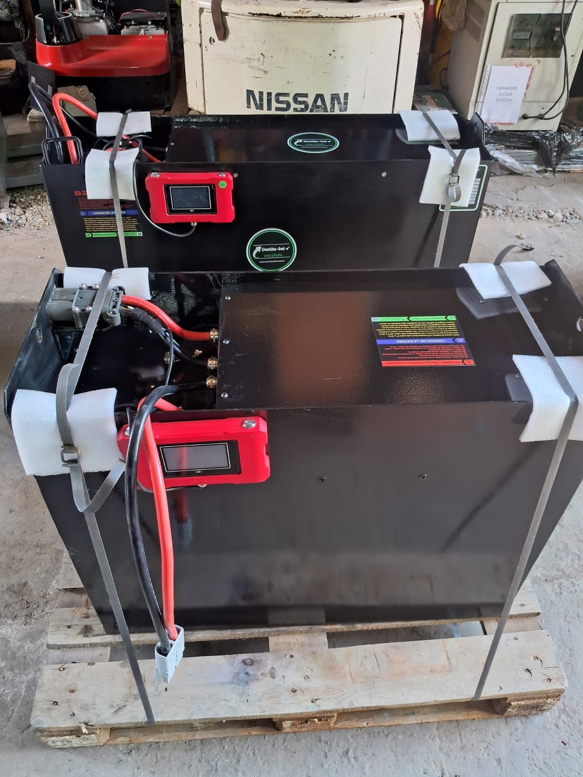 Baterías Nissan negras apiladas sobre un palé de madera, con cables rojos y correas blancas en un taller.