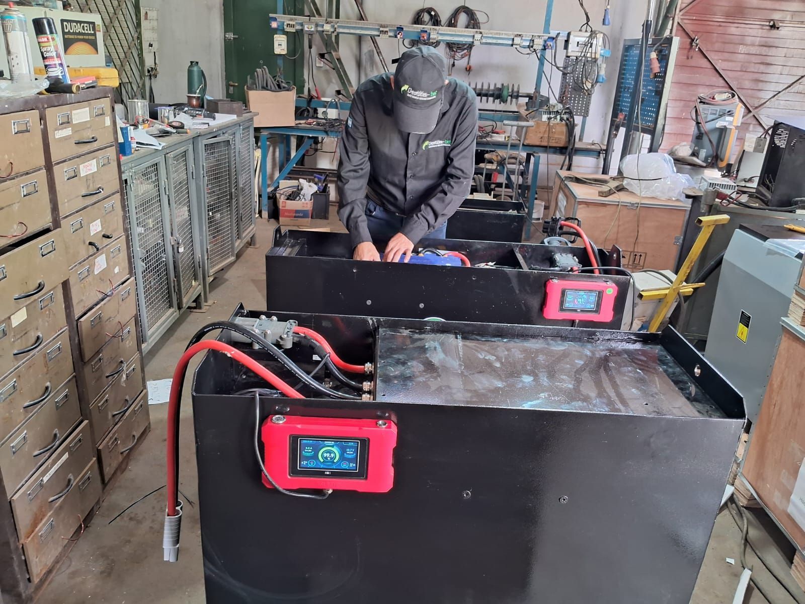 Persona trabajando en una unidad de batería negra en un taller desordenado, con pantallas digitales rojas visibles.