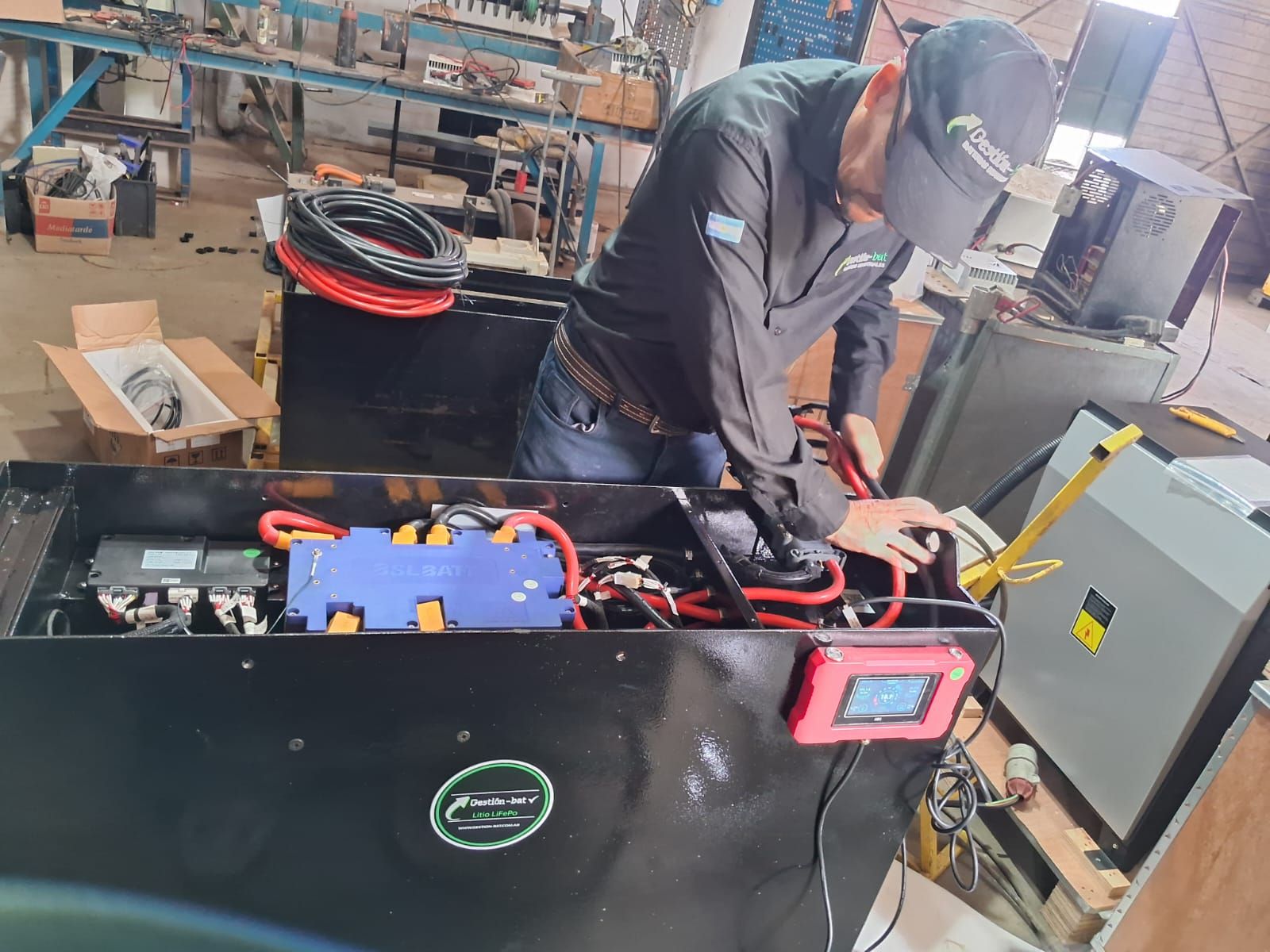 Técnico trabajando en una máquina negra con cables y una pantalla roja en un taller industrial.