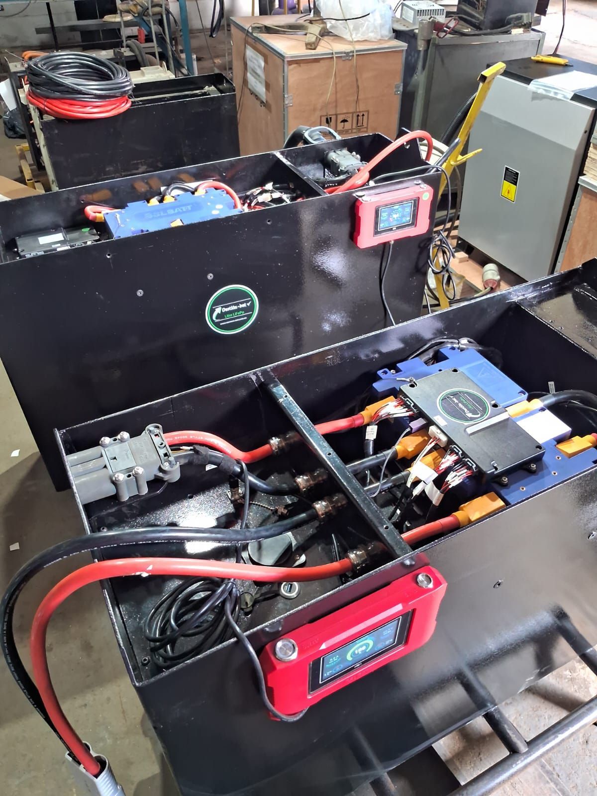 Compartimentos de baterías abiertos con cables, medidores digitales rojos y componentes eléctricos en el suelo de un taller.