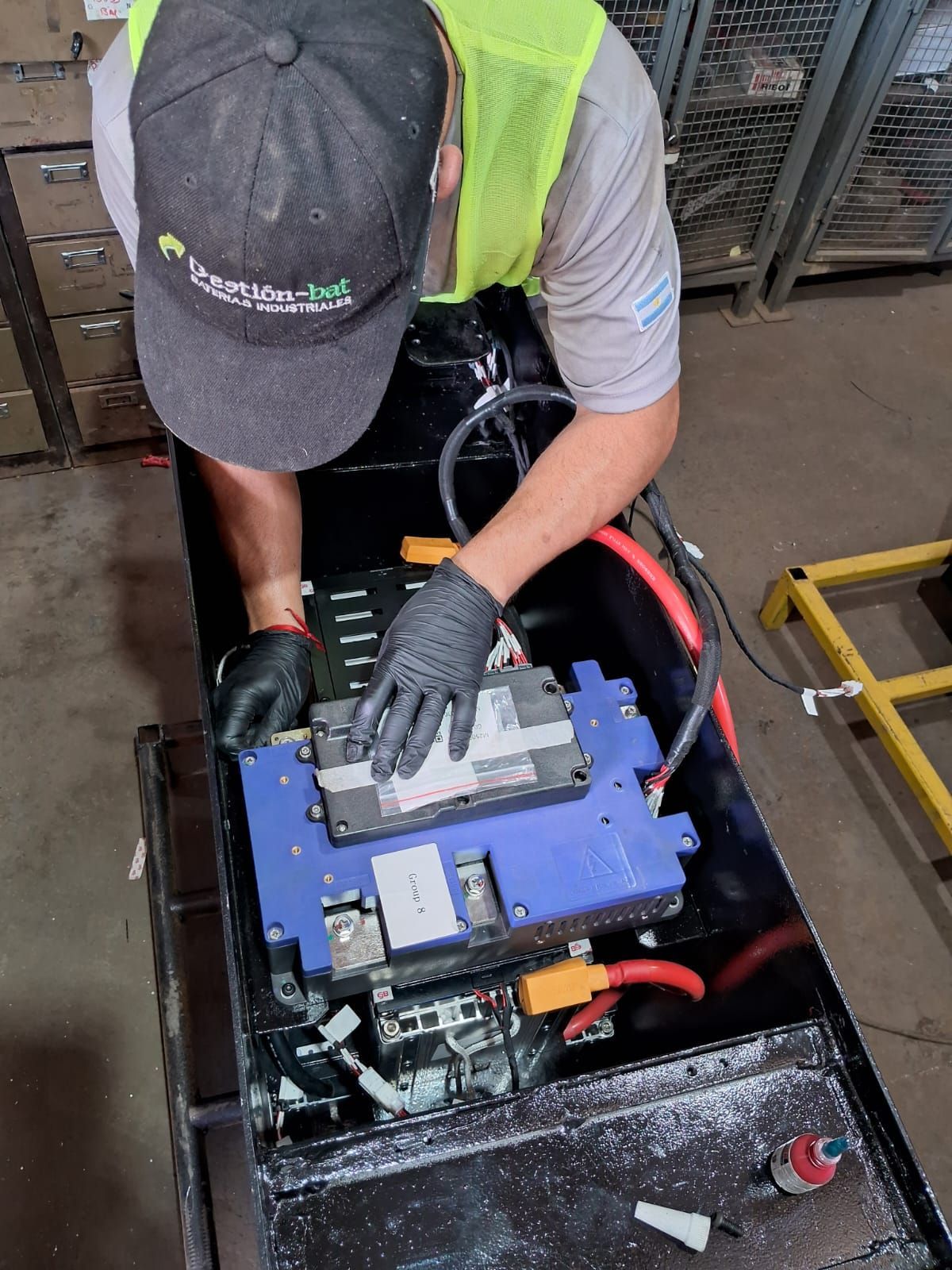 Un trabajador, con chaleco de seguridad y guantes, ajusta una batería azul dentro de un compartimento de equipo negro.