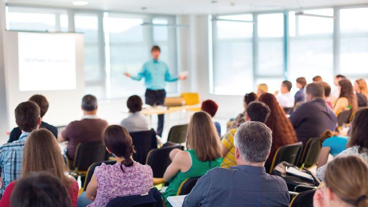 El público, sentado en una luminosa sala de conferencias, observa a un presentador hablar al frente.