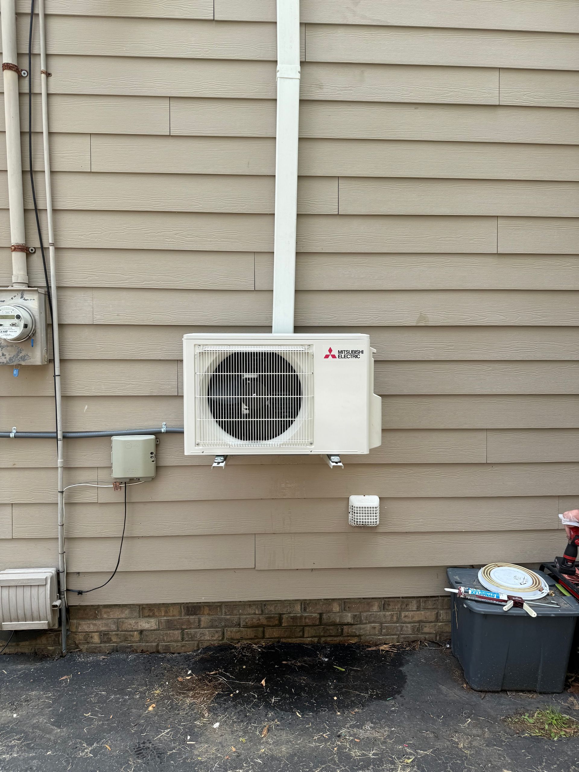 Outdoor wall-mounted air conditioner unit on a beige brick building with pipes and utility boxes nearby.
