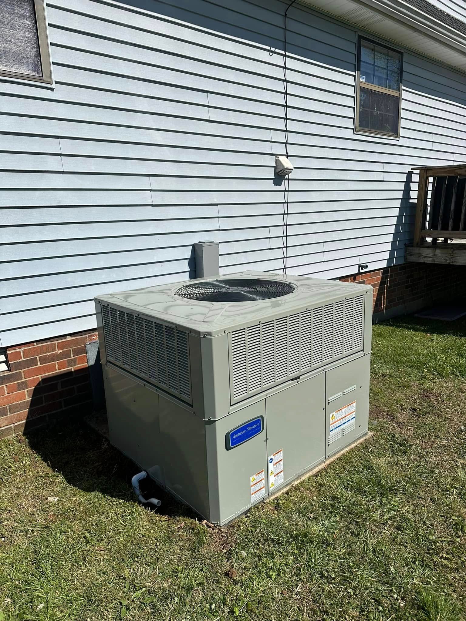 Outdoor HVAC unit beside a house with white siding and a brick foundation, on a grassy lawn.