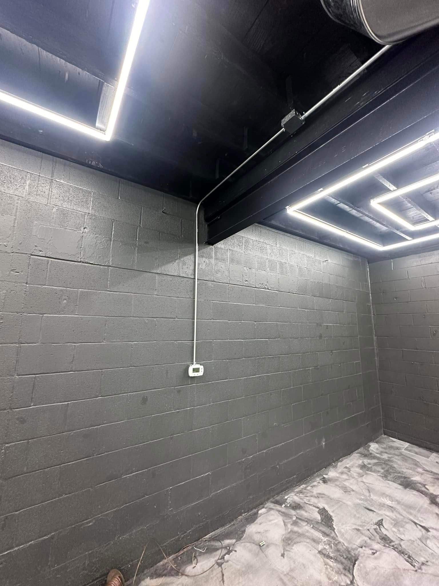 Empty industrial room with dark gray cinderblock walls, exposed ceiling lights, and a concrete floor.
