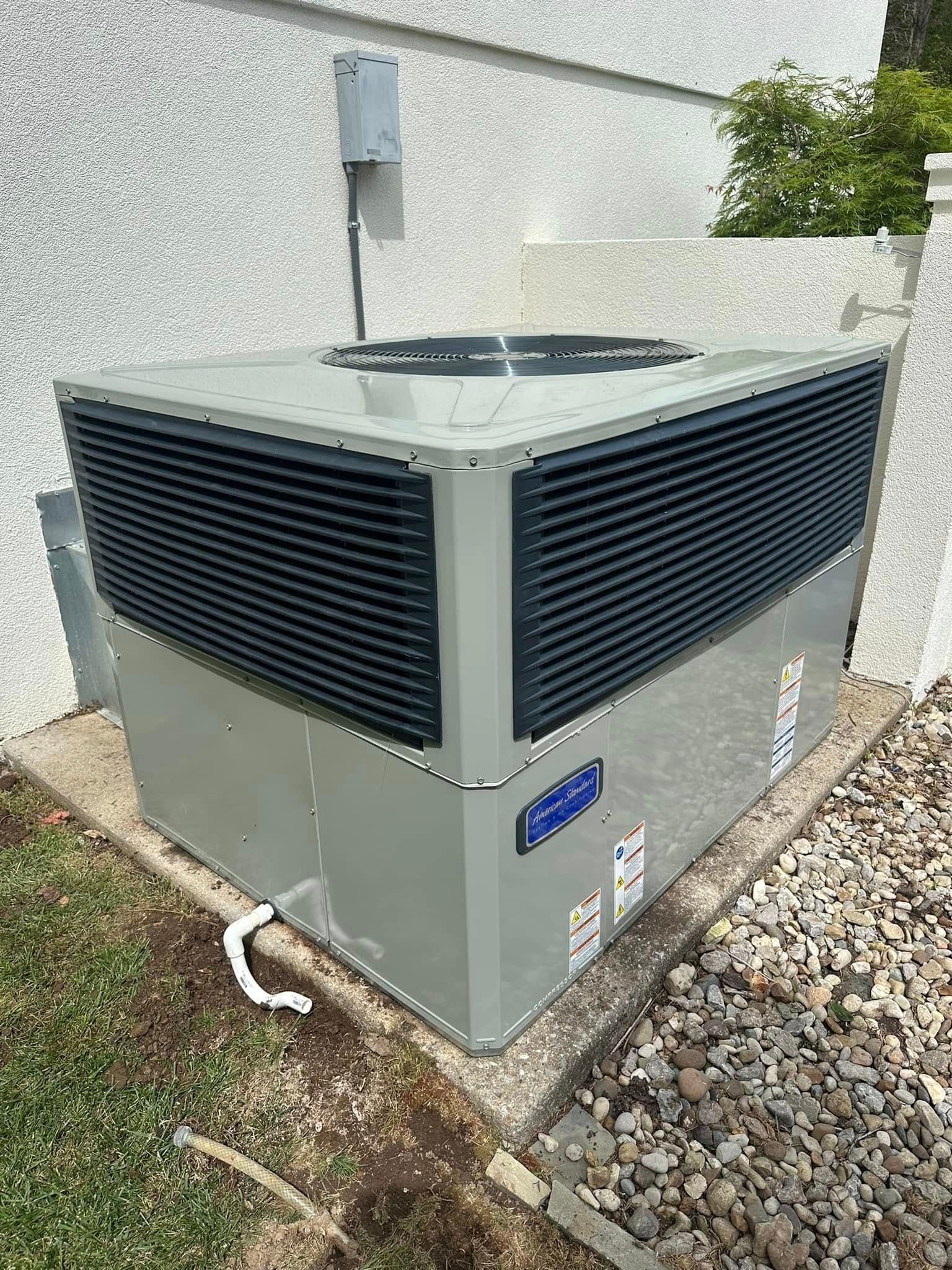 Outdoor HVAC condenser unit beside a building wall, with dark vents and gray casing.