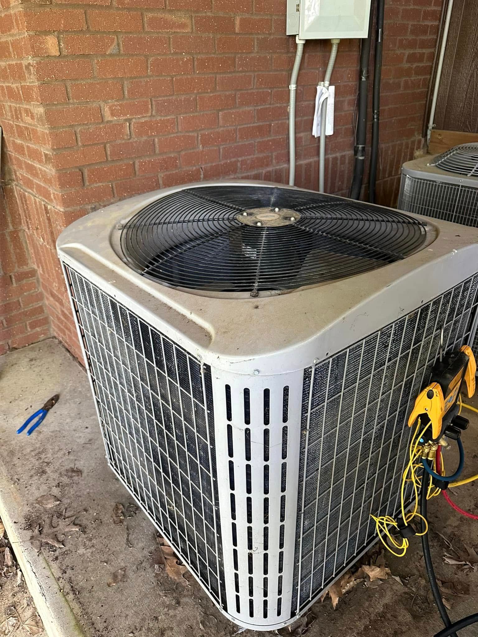 Outdoor air conditioner unit beside a brick wall, with tools and hoses nearby.