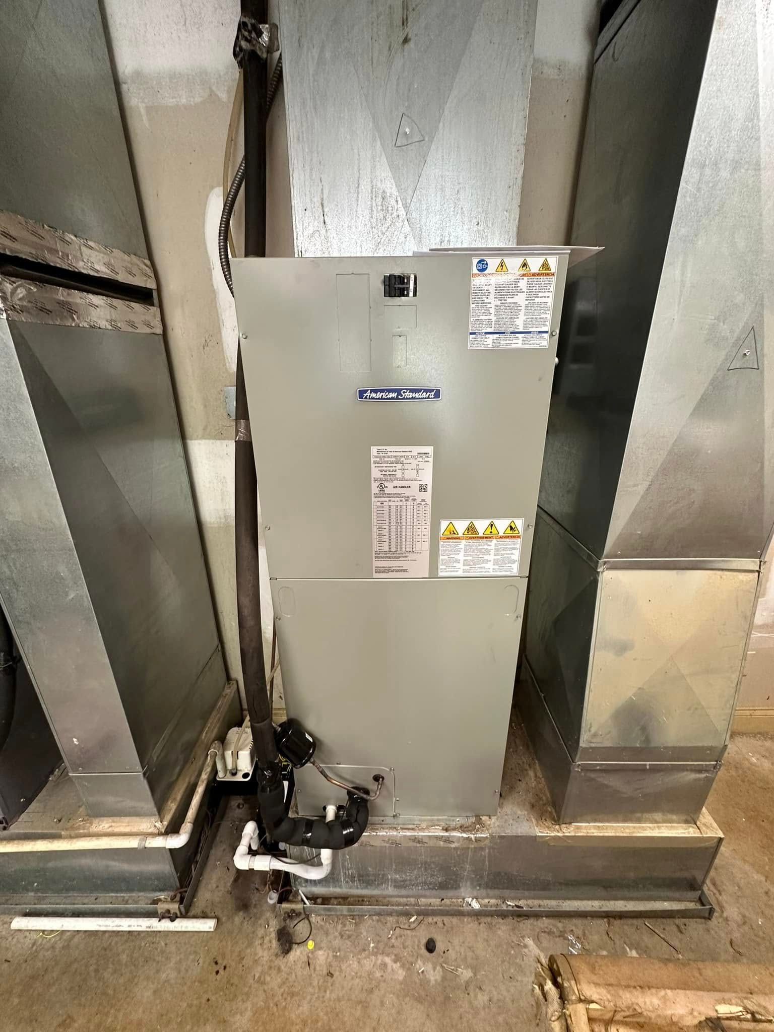 Metal HVAC unit in a mechanical room, with labels and piping at the base between ductwork.