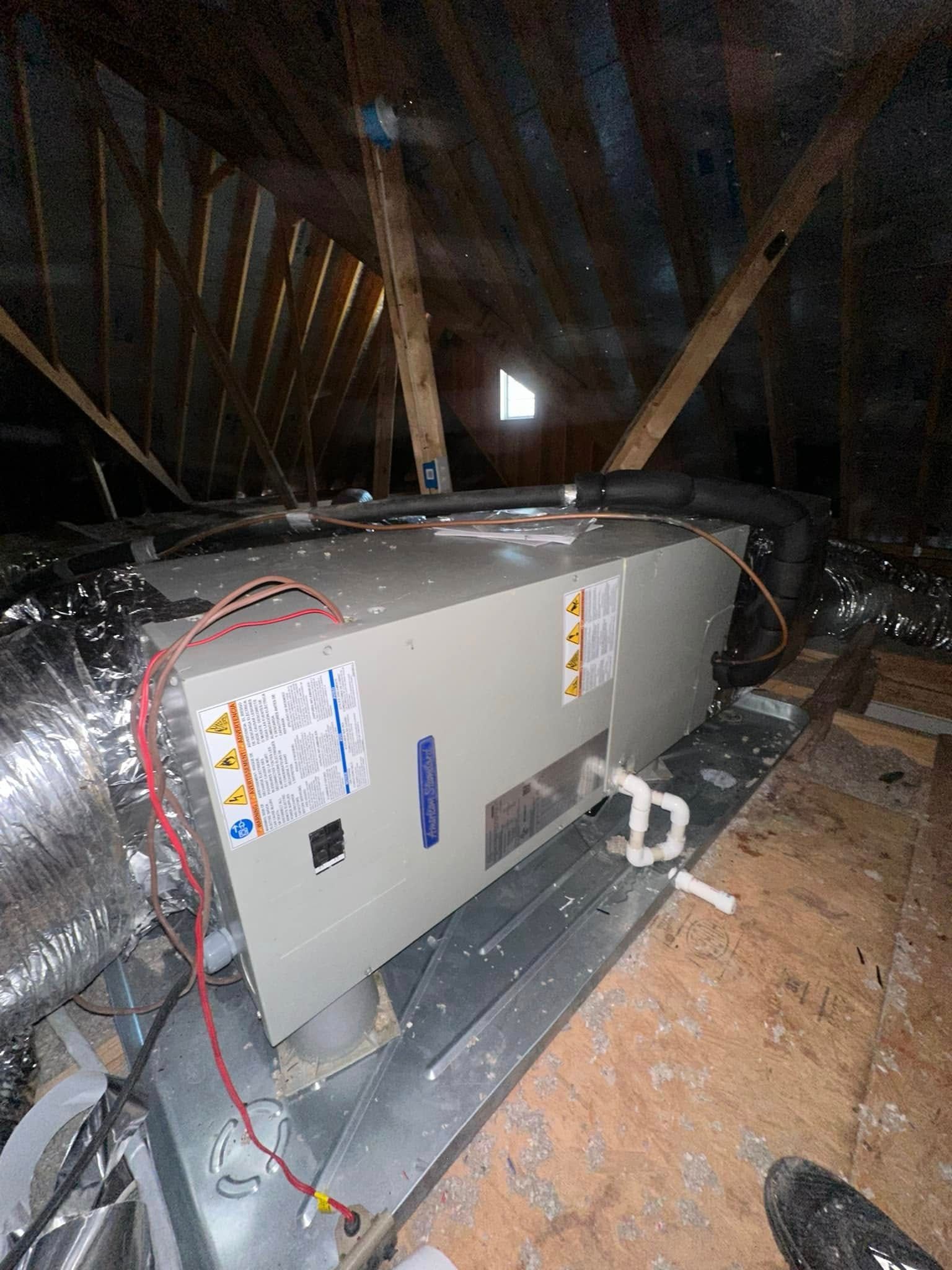 Gray HVAC unit in an attic, with ducting and wires on the floor and exposed rafters overhead