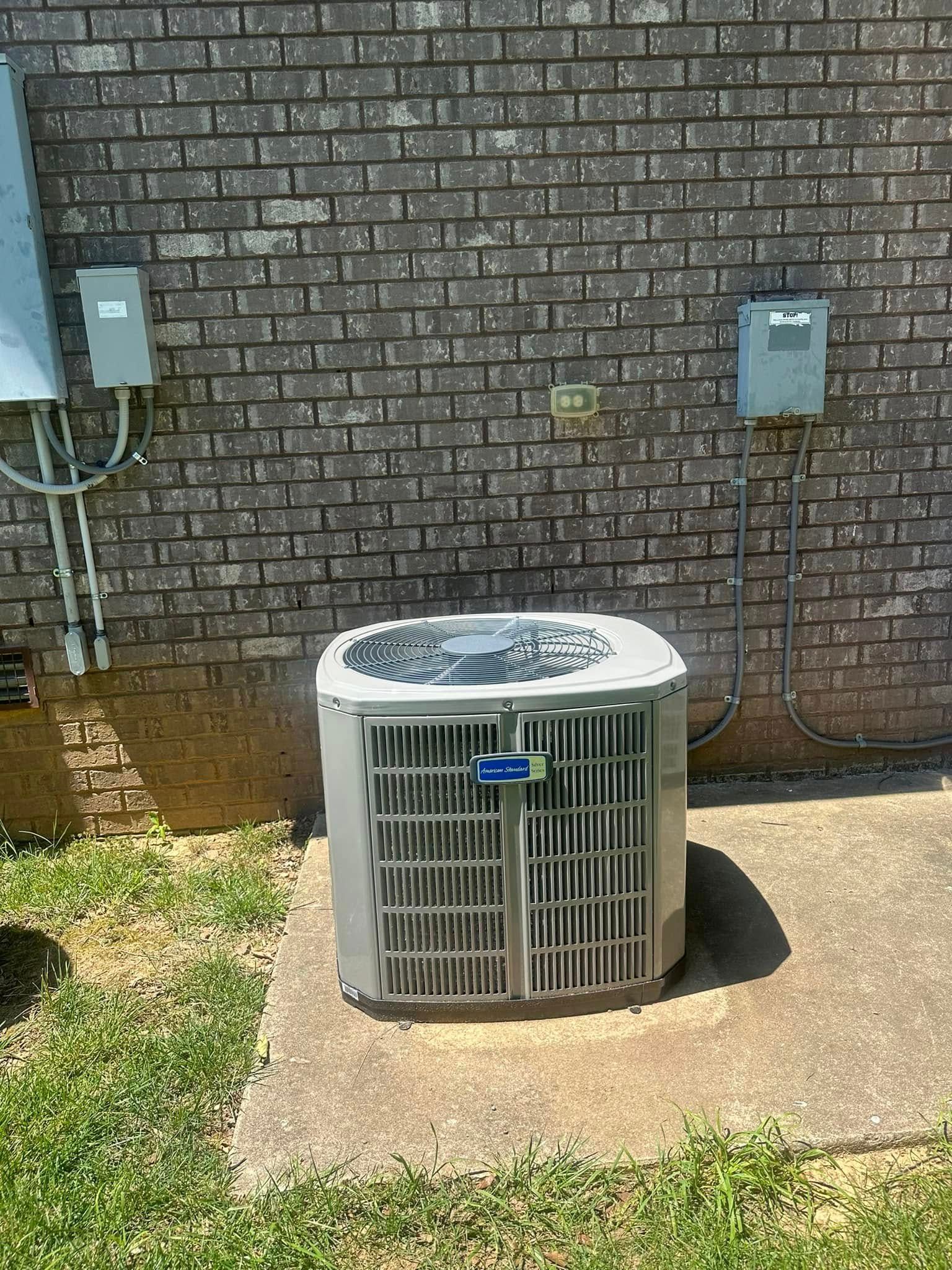 Outdoor air conditioner unit beside a brick wall with utility boxes and a concrete pad