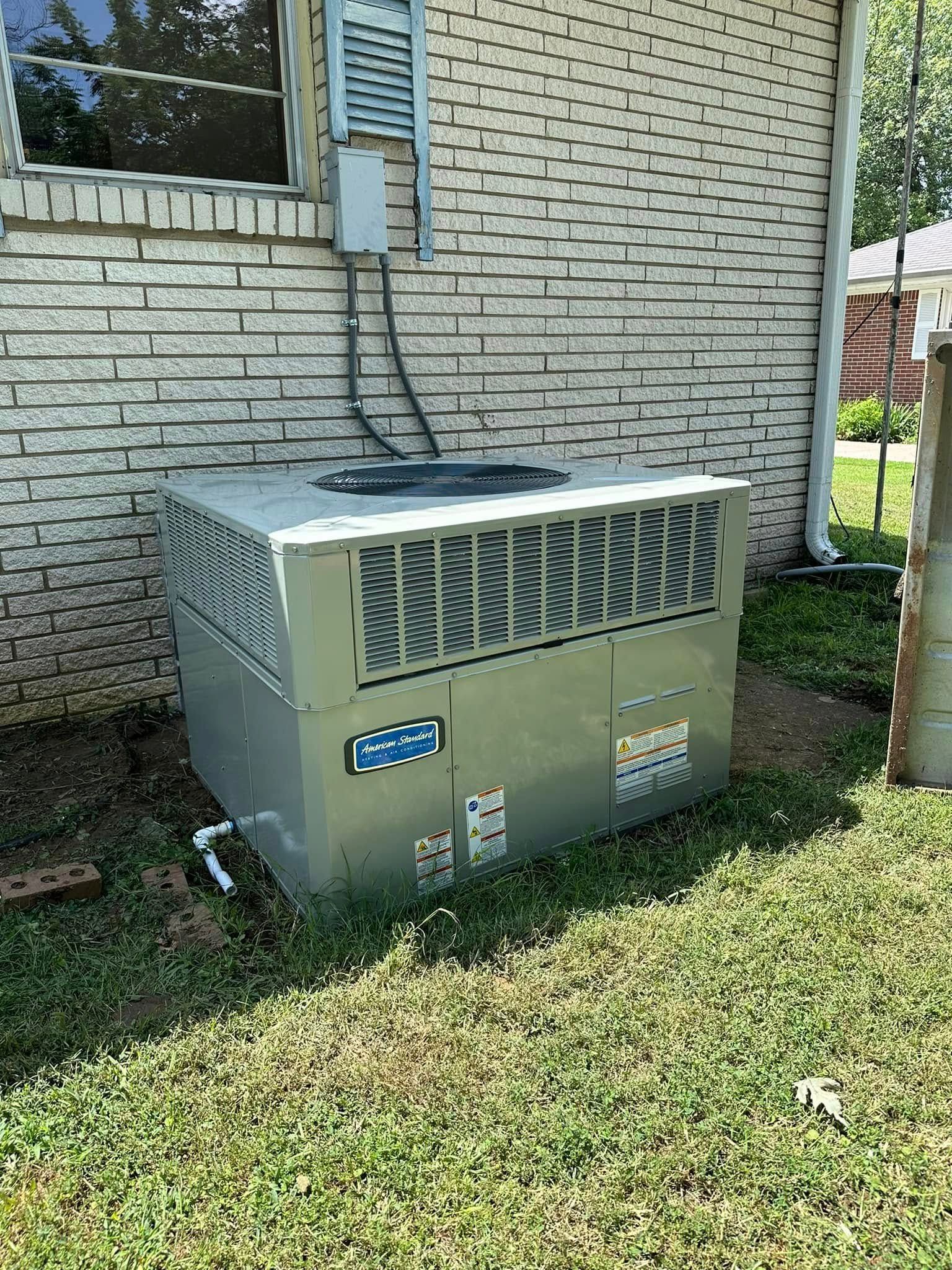 Outdoor HVAC unit beside a brick house, with pipes and grass in the foreground.