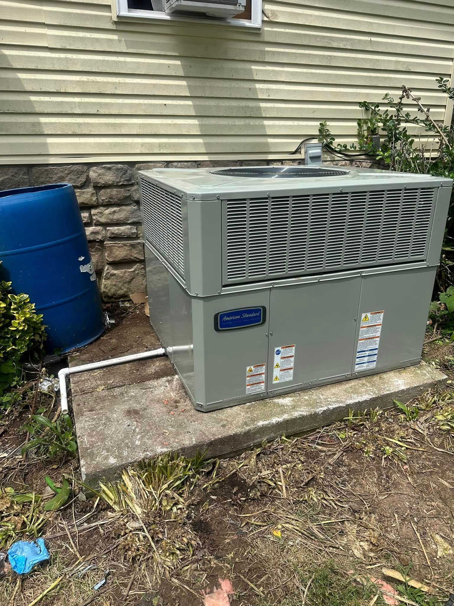 Outdoor HVAC unit beside a beige house, with a blue barrel nearby and a small garden bed.