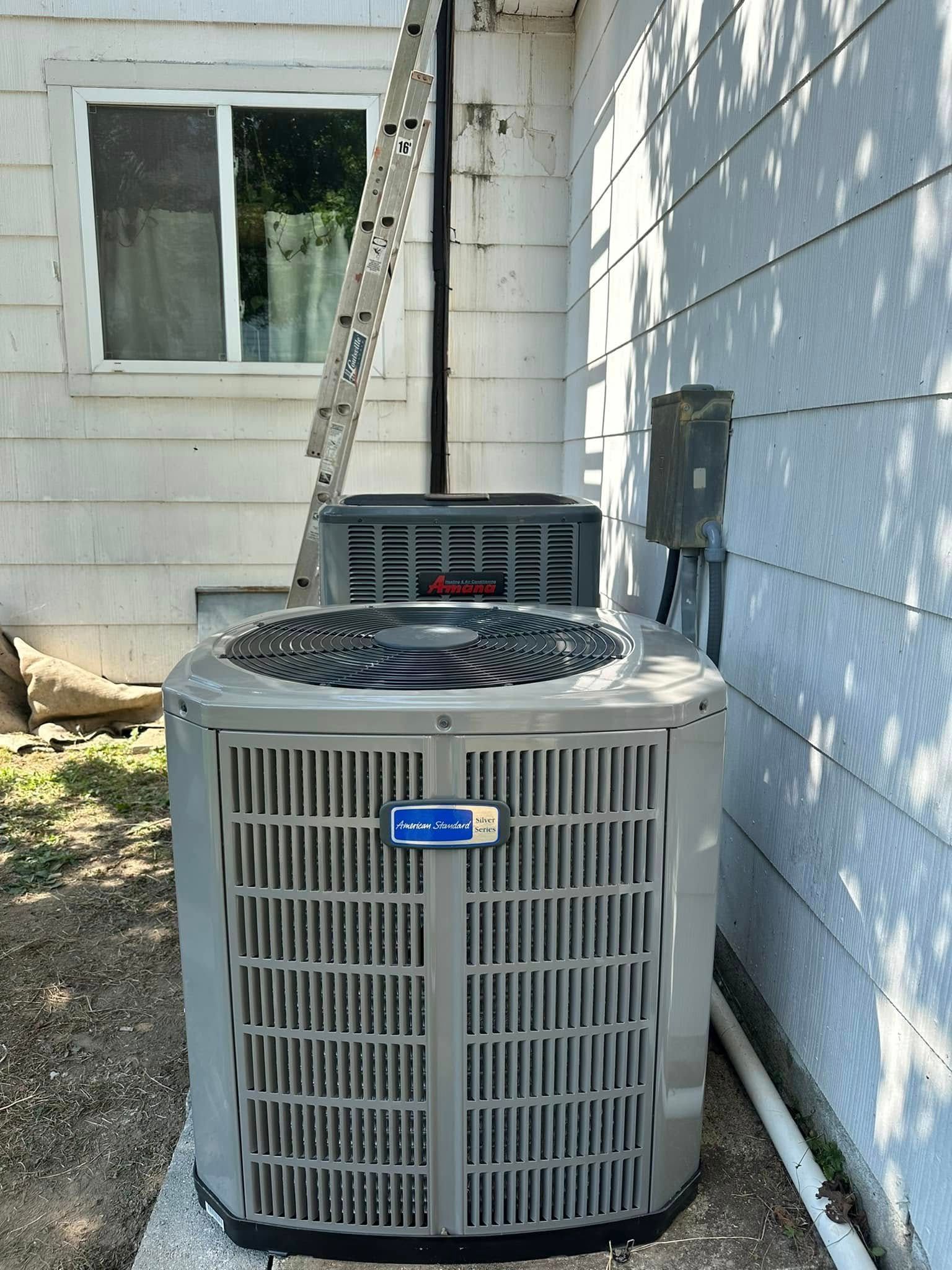 Outdoor air conditioning unit beside a white house wall and window, with a ladder in back.