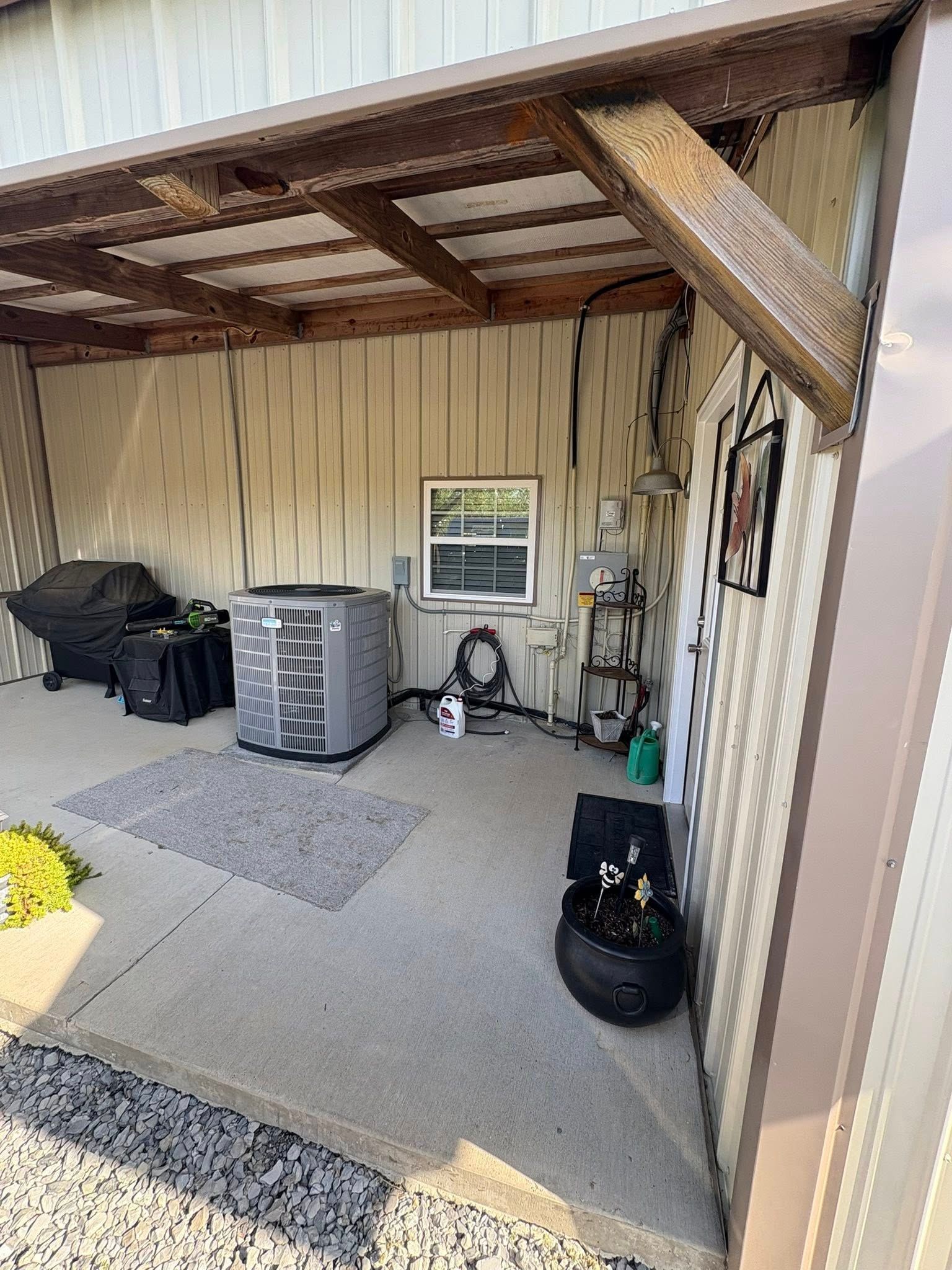 Covered patio with a large grill, storage bin, and black bag beside a window and tan siding.