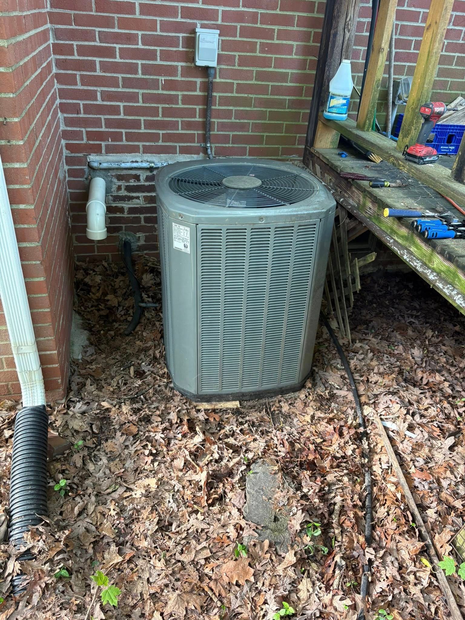 Outdoor air conditioner unit beside a brick wall, surrounded by leaves and a workbench.