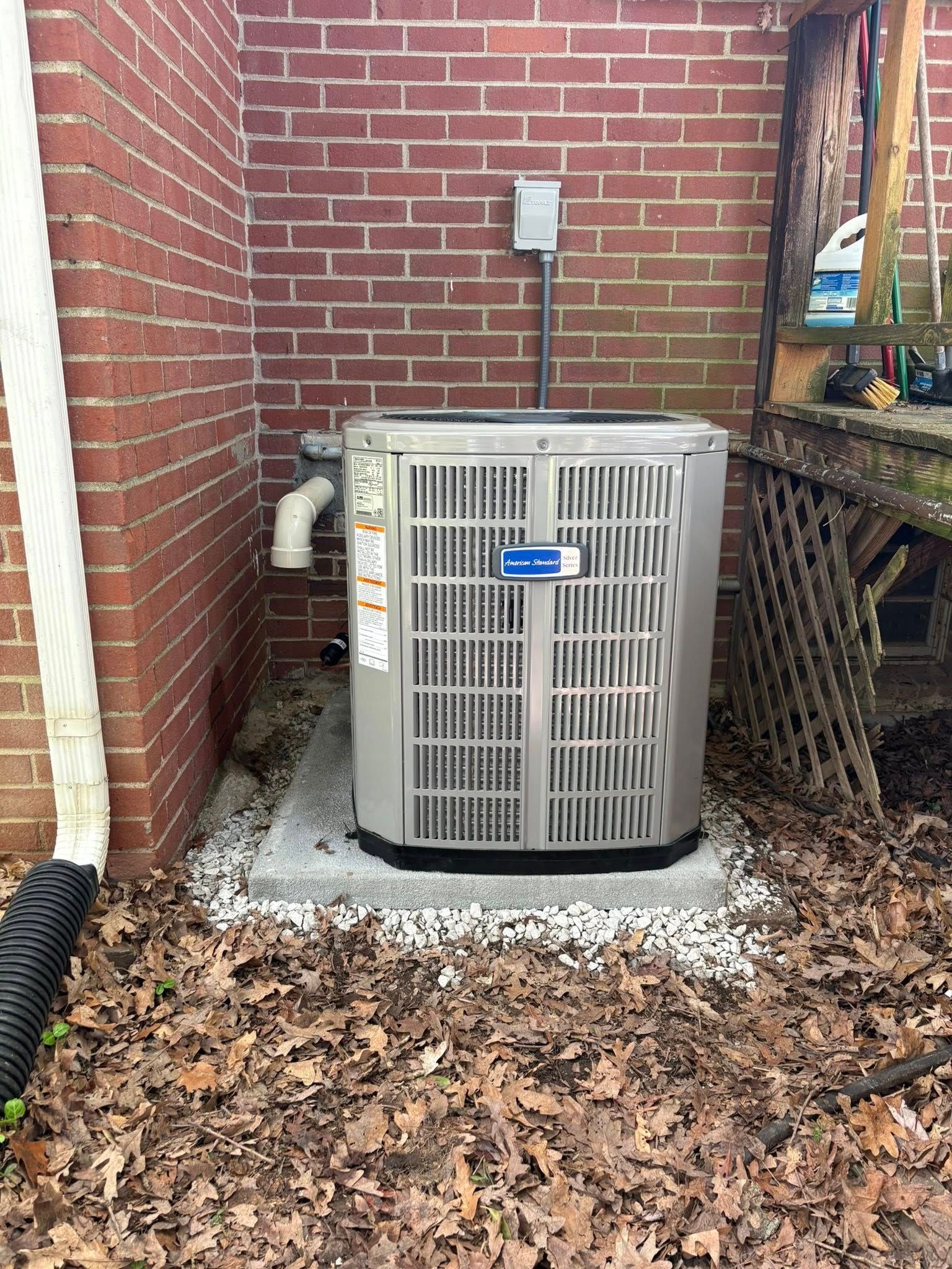 Outdoor HVAC unit beside a brick house wall, surrounded by fallen leaves and a concrete pad