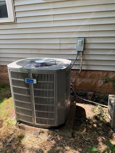 Outdoor air conditioner unit beside a house wall, with electrical conduit and dry grass nearby